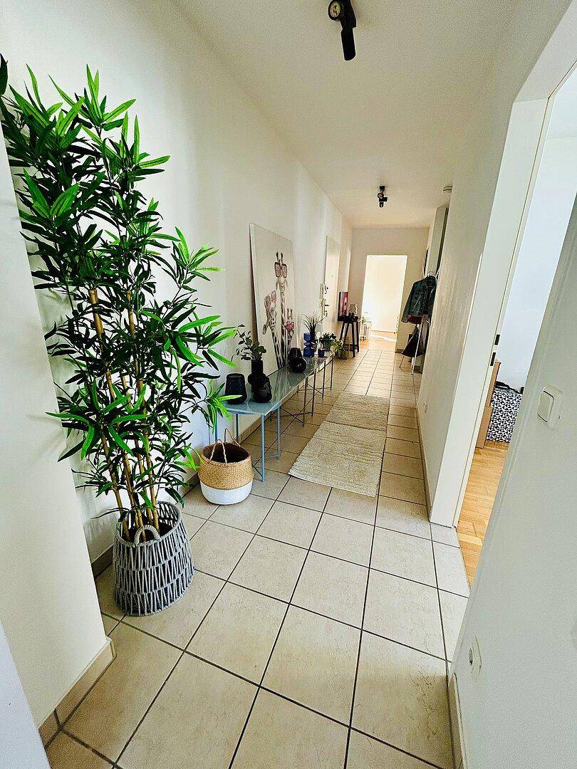 Langer, heller Flur mit Fliesenboden, einer großen Pflanze und moderner Wanddekoration.