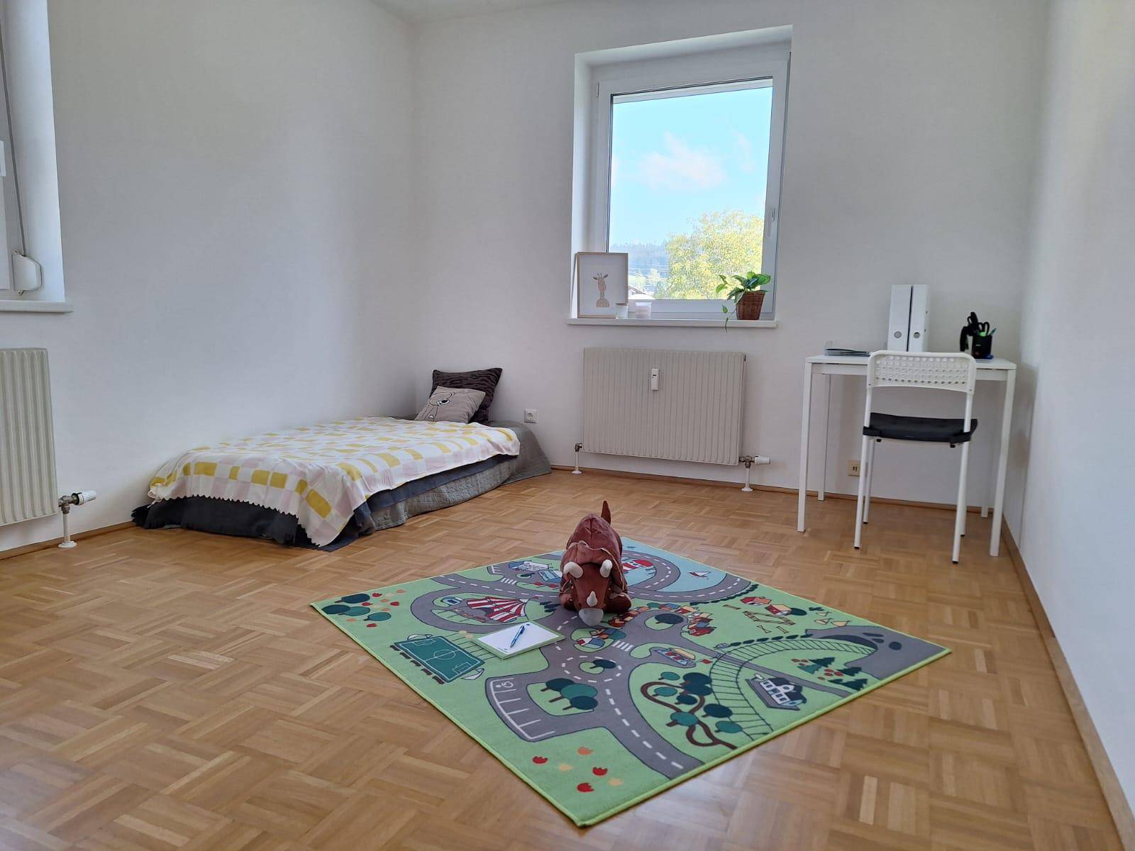 Kinderzimmer mit Parkettboden, einem Bett, Schreibtisch und einem bunten Spielteppich.