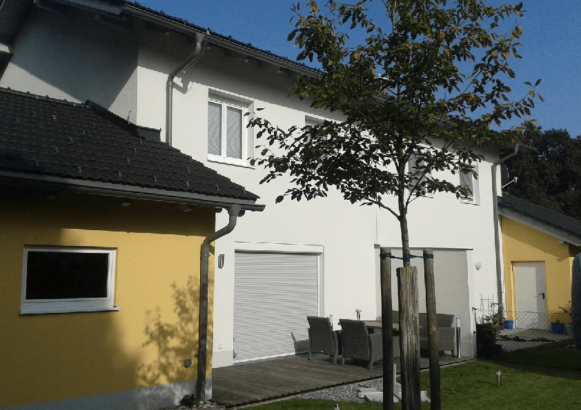 Moderne Hausfassade mit weißem Putz, einer Holzterrasse und gepflegtem Gartenbereich.