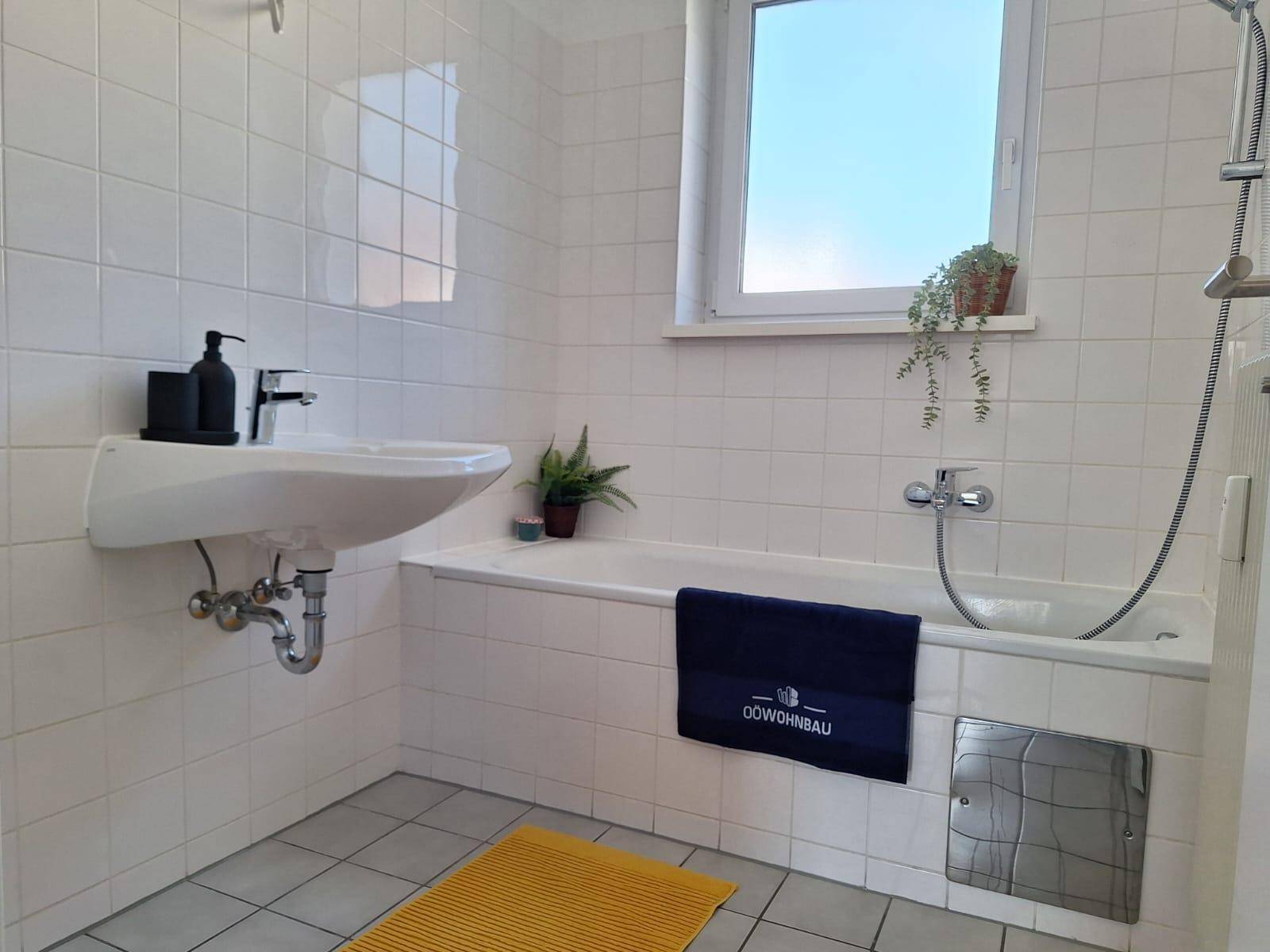 Badezimmer mit Badewanne, Waschbecken, Fenster und weißen Fliesen an den Wänden.