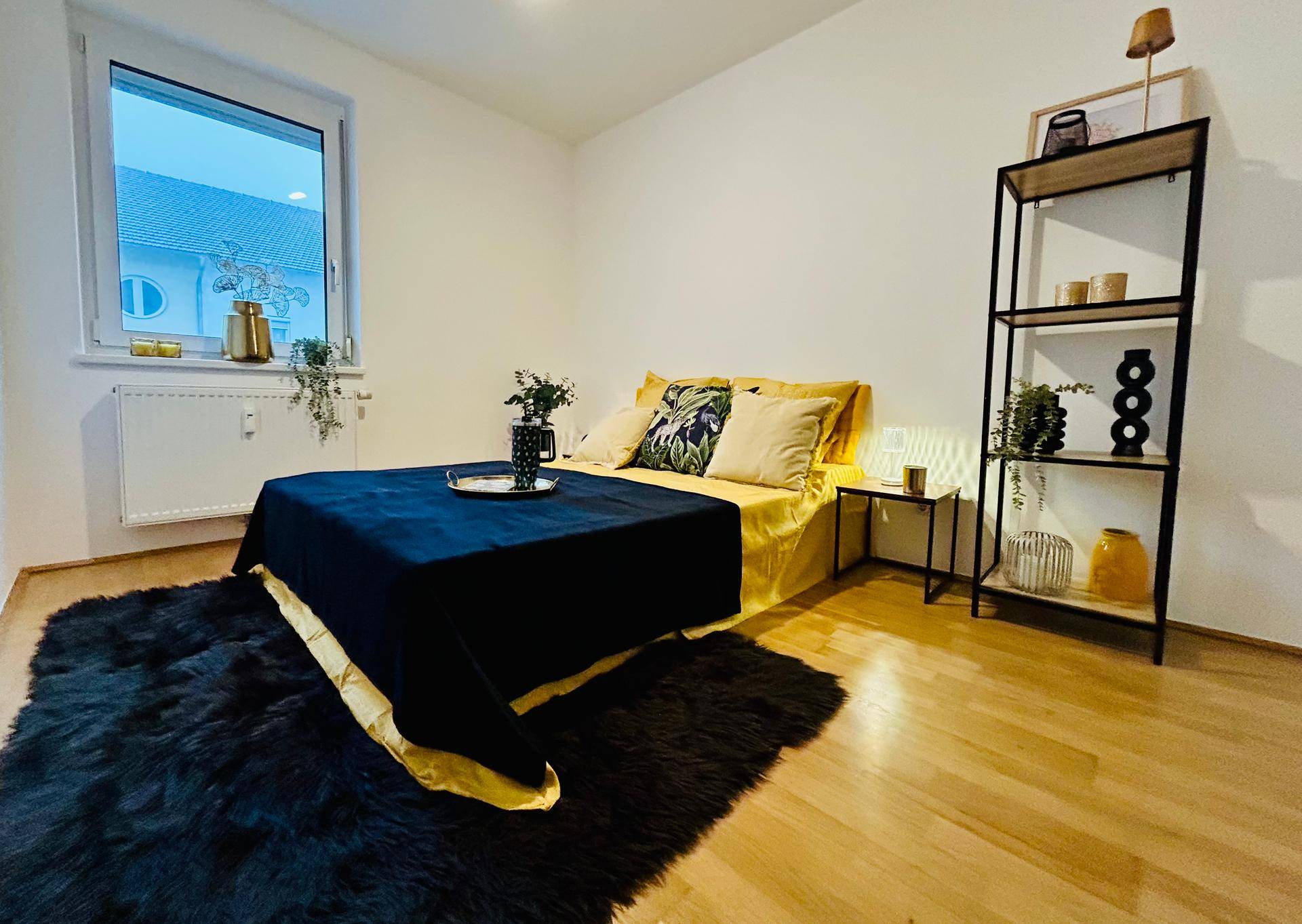 Helles Schlafzimmer mit Holzboden, großem Bett, schwarzem Teppich und einem modernen Regal.