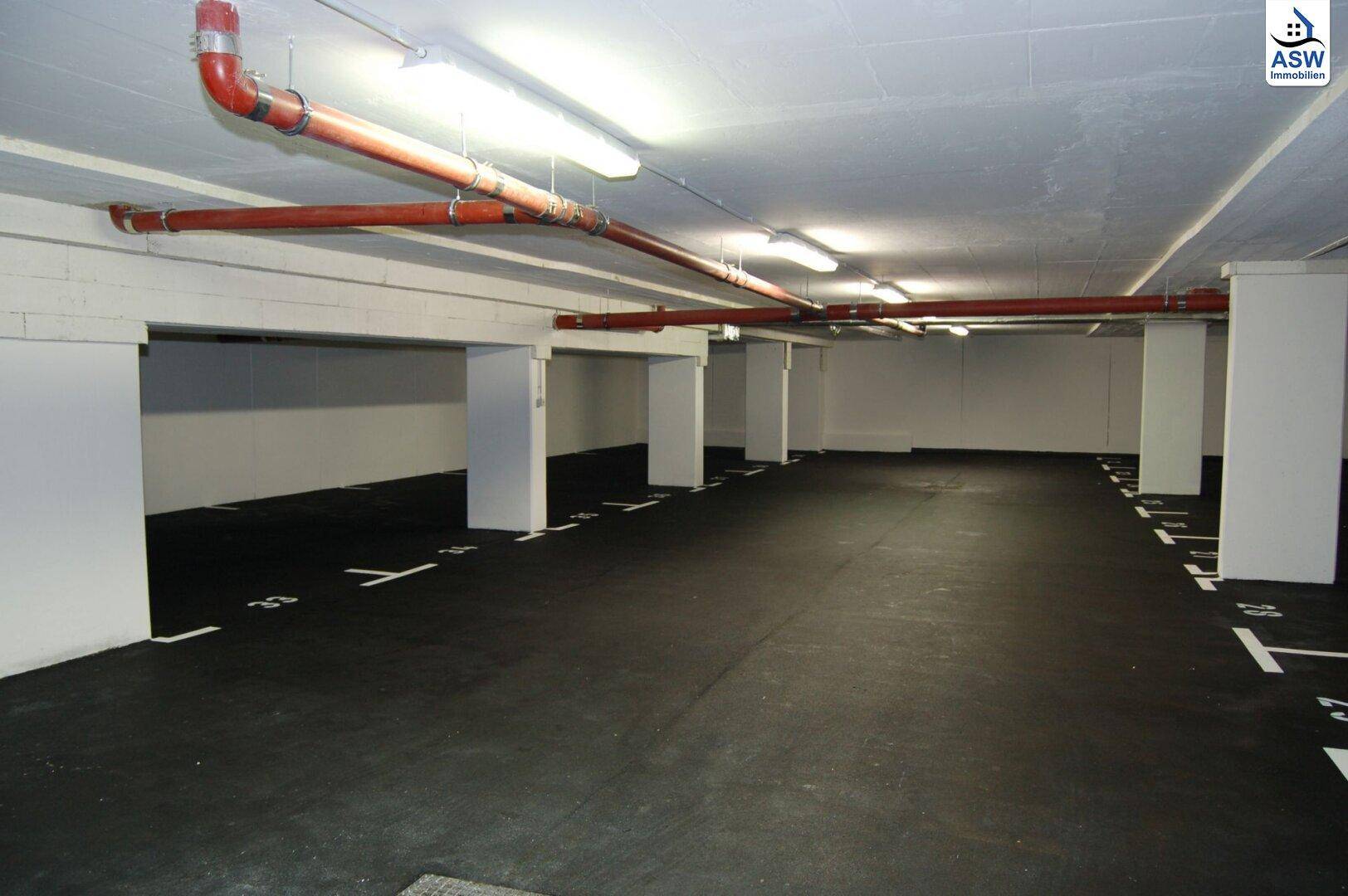 Helle Tiefgarage mit zahlreichen Stellplätzen, weißen Säulen und sichtbaren Versorgungsleitungen an der Decke.