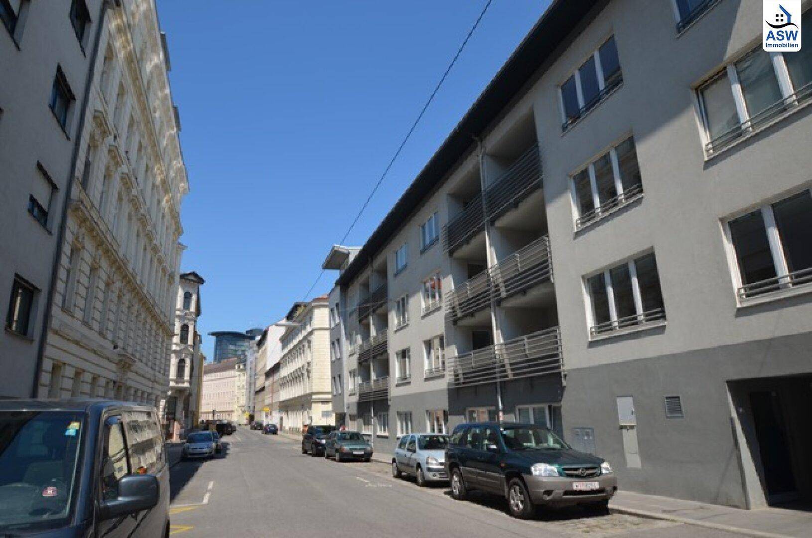 Städtische Straße mit parkenden Autos und Gebäuden unter blauem Himmel.