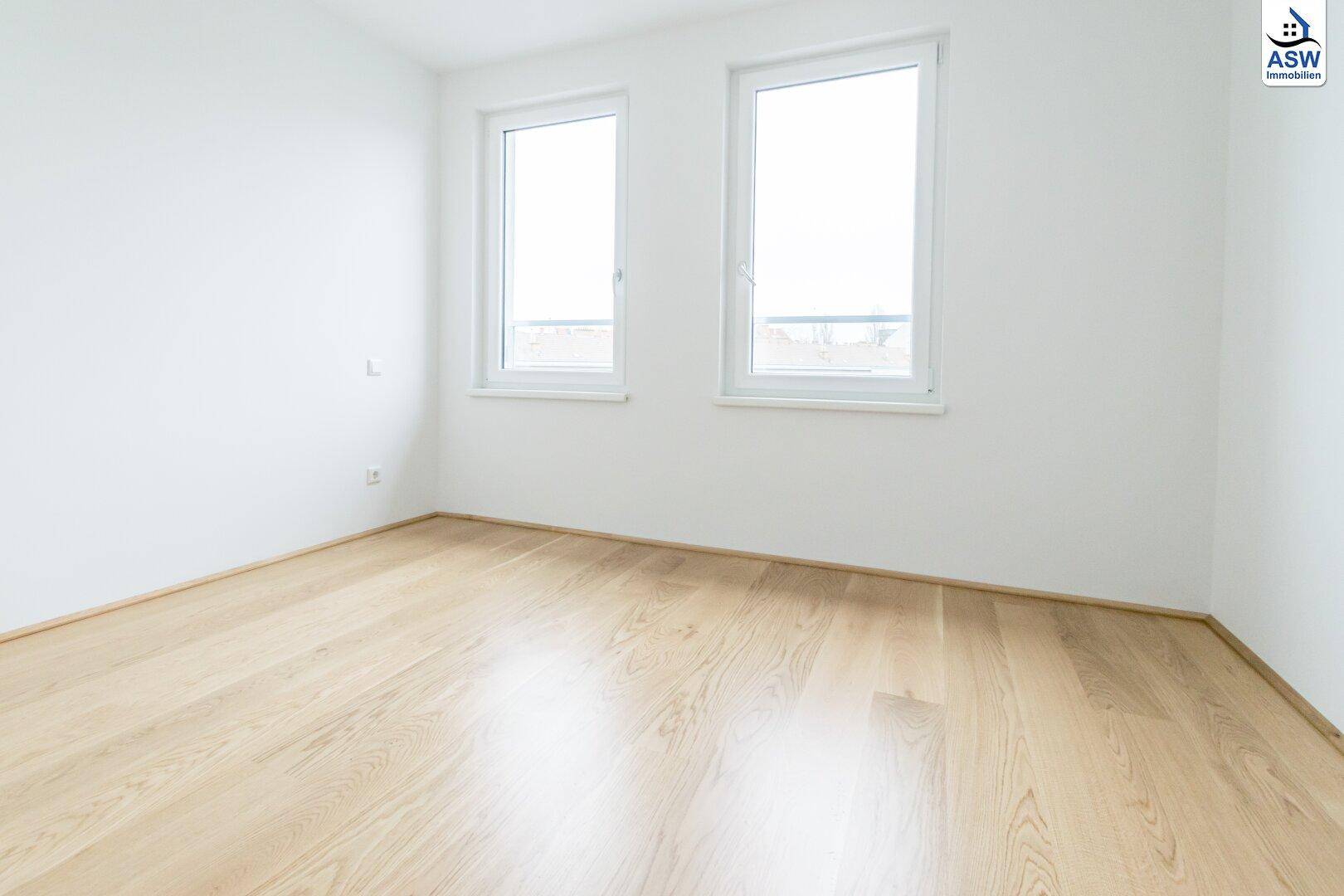 Helles, leeres Zimmer mit zwei großen Fenstern und schönem Holzparkettboden.