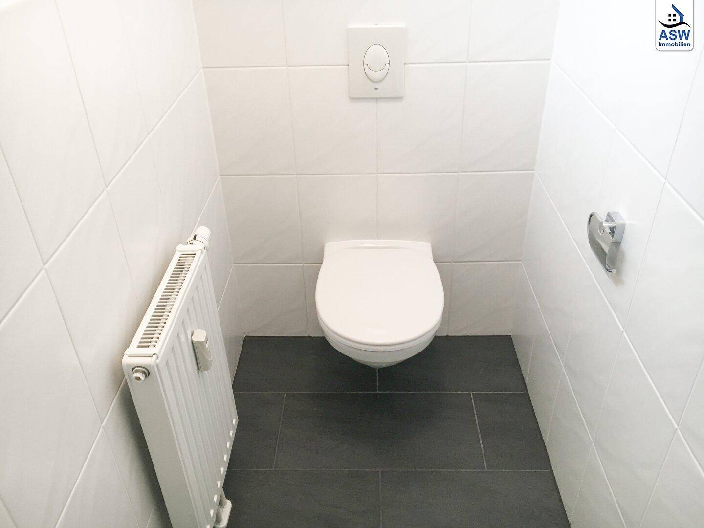 Separate Toilette mit weißen Wandfliesen, dunklem Boden und wandhängendem WC.