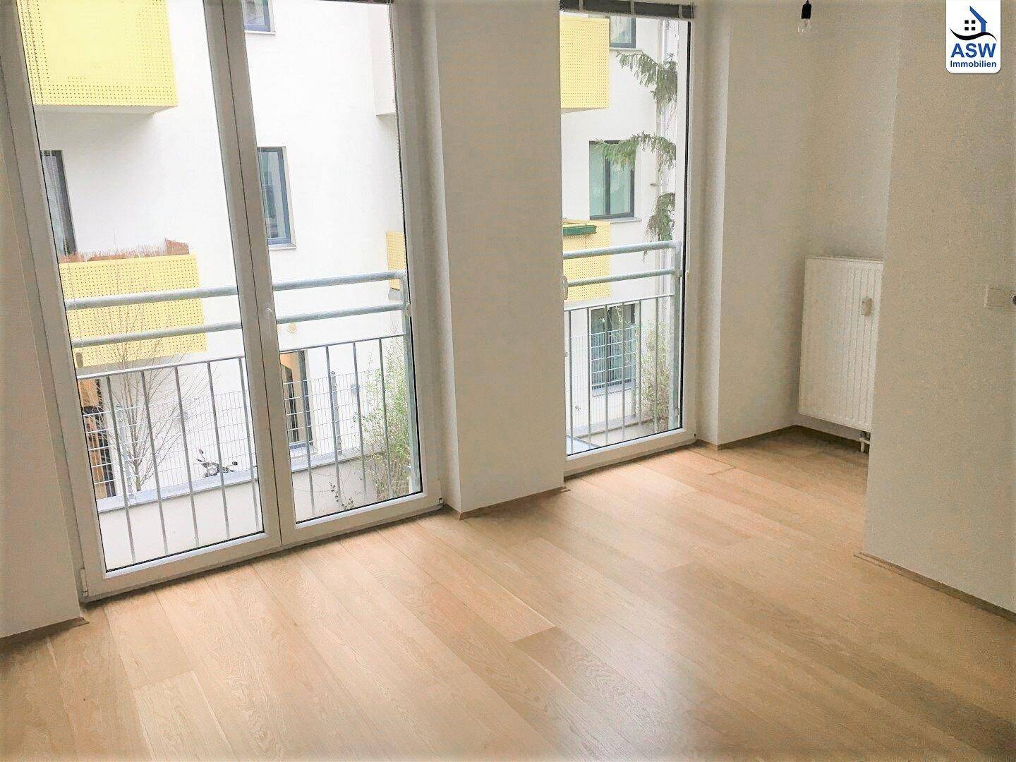 Heller Wohnraum mit Holzboden, weißen Wänden und Zugang zu einem Balkon mit Innenhofblick.