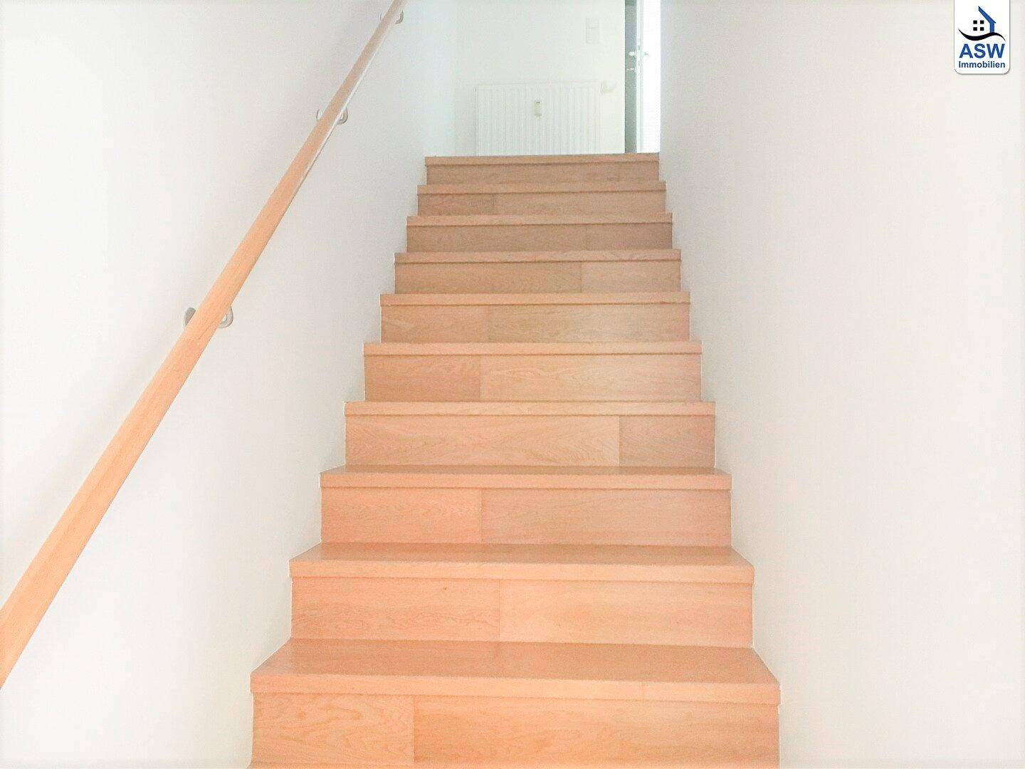 Helle Holztreppe mit schlichtem Holzgeländer und weißen Wänden führt in die obere Etage.