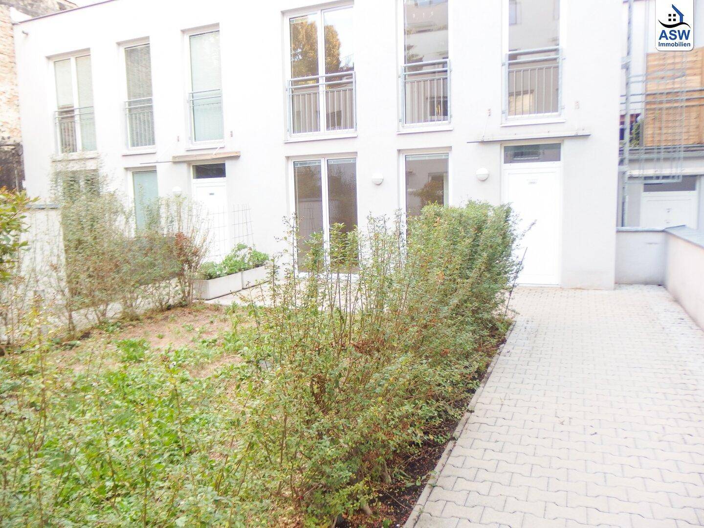 Außenansicht eines modernen Gebäudes mit Gartenbereich, gepflastertem Weg und Balkonen.