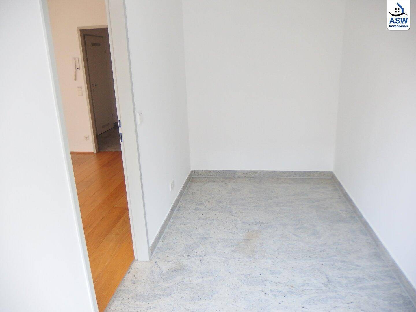 Kleiner Flurbereich mit grauen Fliesen und weiß gestrichenen Wänden, angrenzend an einen Raum mit Holzboden.