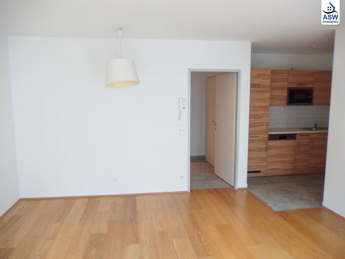 Heller Wohnbereich mit Holzboden, weißer Wand und Blick auf die angrenzende Küche.