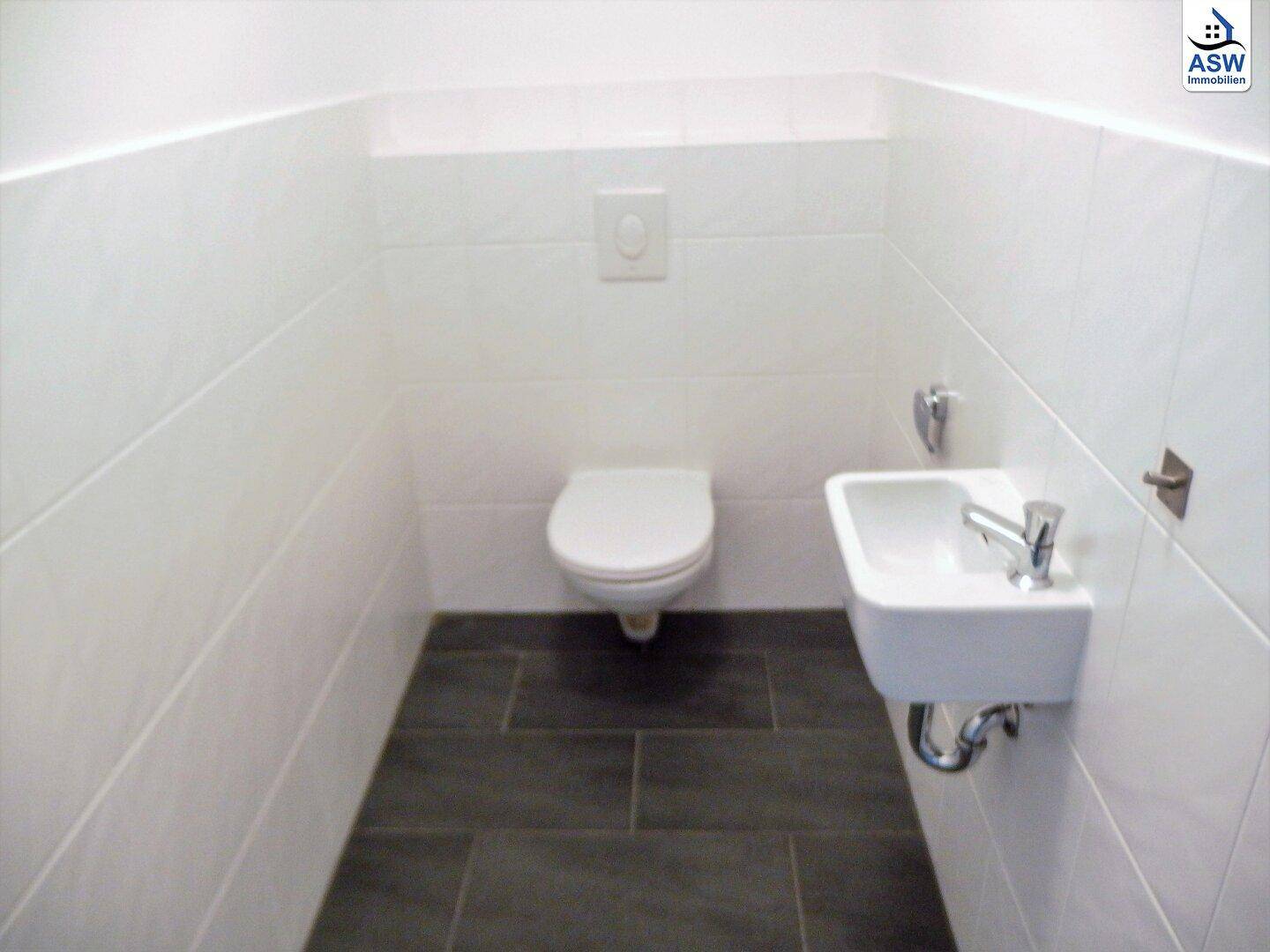 Gäste-WC mit weißen Wandfliesen, dunklem Boden, wandhängendem WC und kleinem Handwaschbecken.
