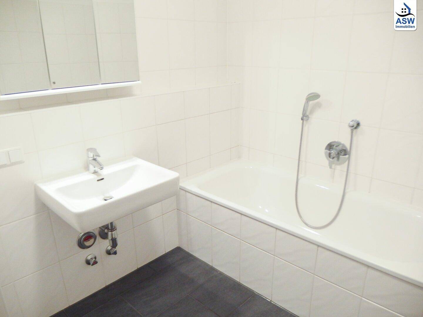Badezimmer mit Badewanne, Waschbecken und weißen Wandfliesen, ergänzt durch dunkle Bodenfliesen.