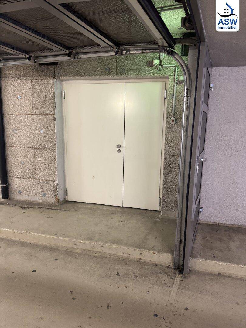 Eingang zu einer Lagereinheit in einer Tiefgarage mit weißen Doppeltüren und rauen Betonwänden.