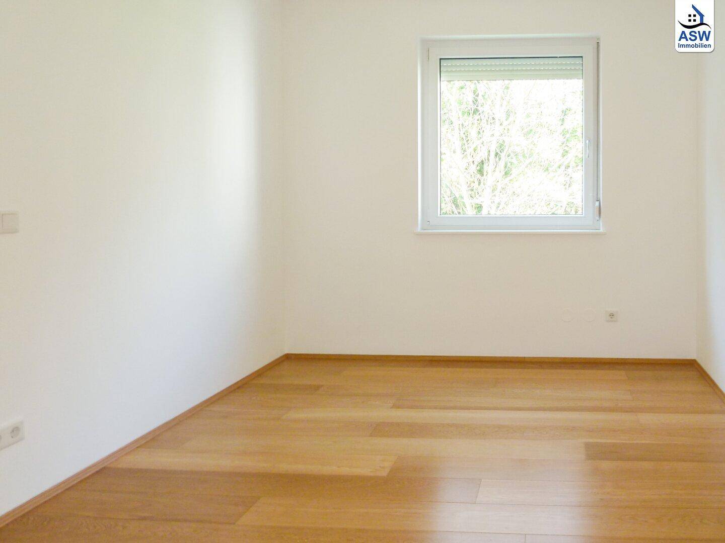 Heller Raum mit Holzboden und einem Fenster, das einen angenehmen Blick ins Grüne bietet.