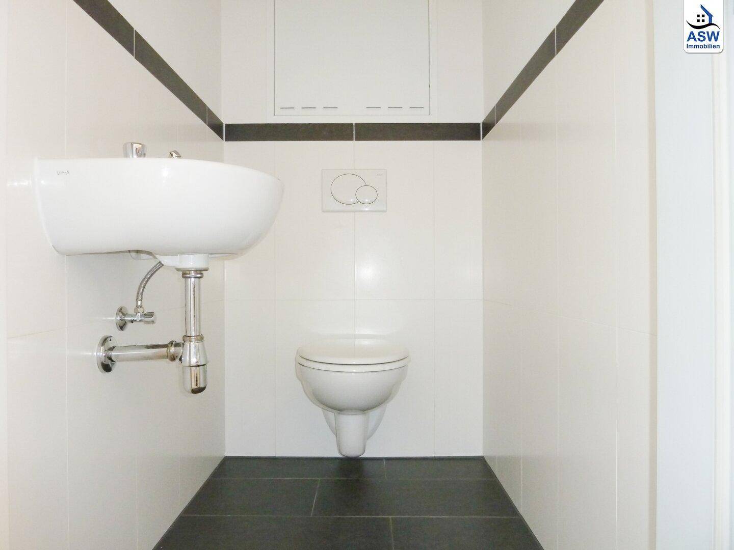 Separates WC mit wandhängender Toilette, Waschbecken und kontrastierenden weißen und dunklen Fliesen.