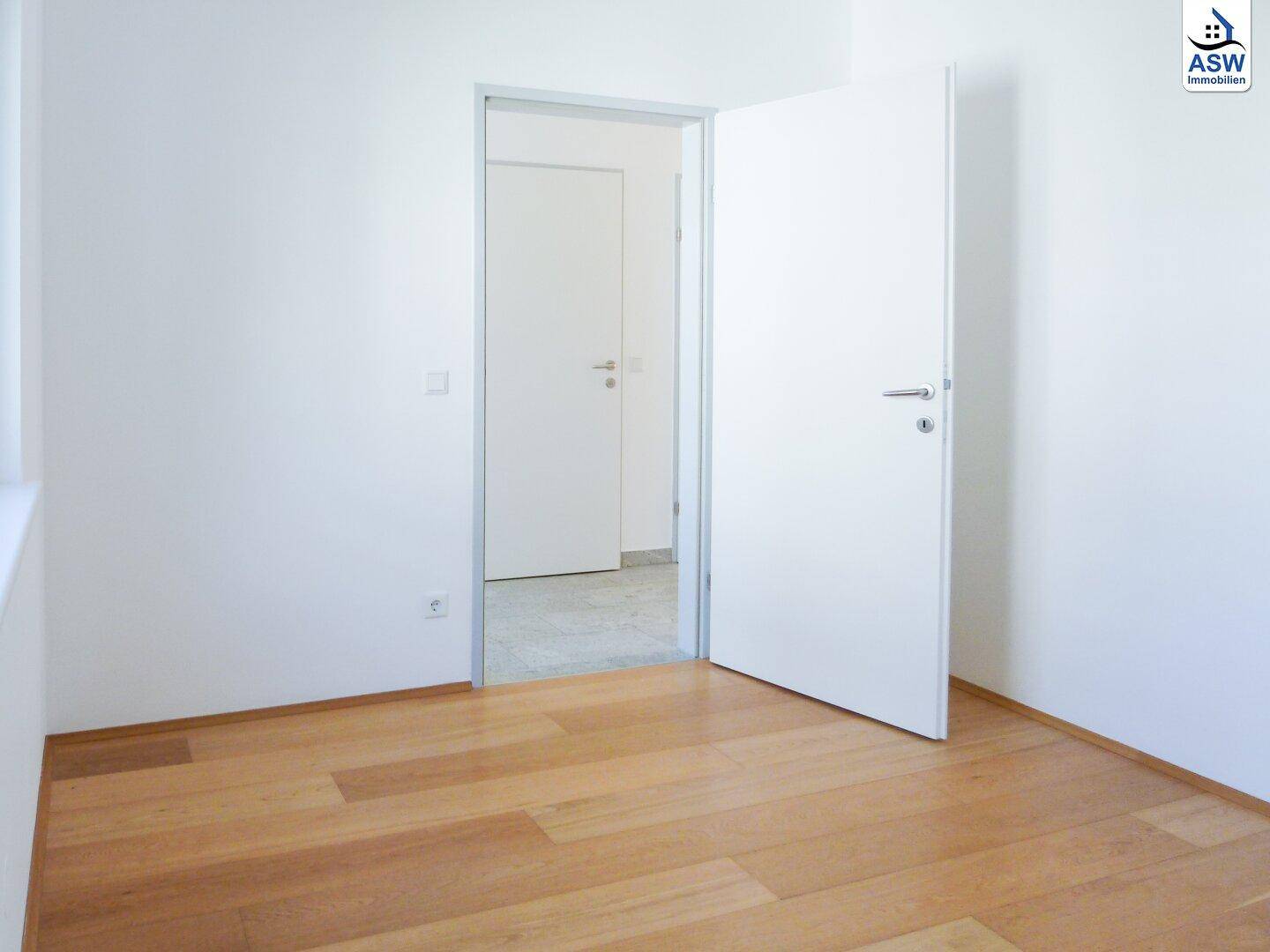 Leerer Raum mit Holzboden und einer offenen Tür, die zu einem angrenzenden Bereich führt.