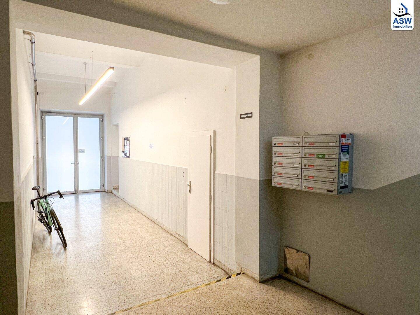 Langer, heller Hausflur mit Terrazzoboden, Briefkästen und einem Fahrrad am Ende.