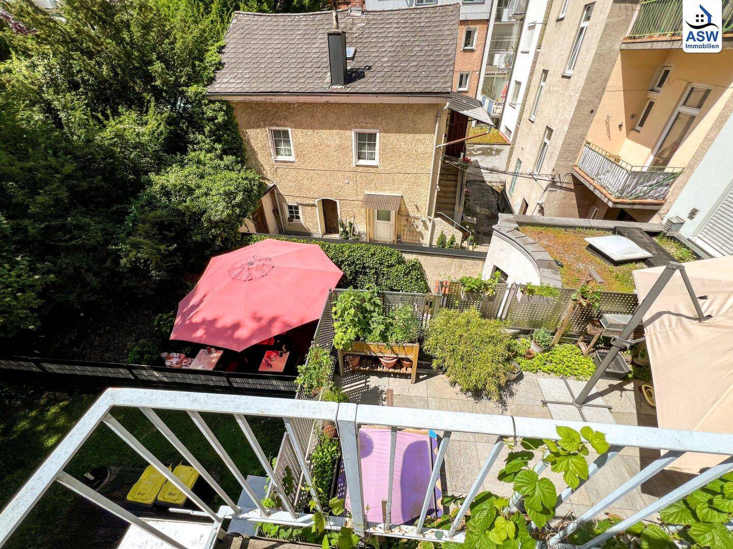Blick von einem Balkon auf einen grünen Hinterhof mit Sonnenschirm und einem kleinen Haus.