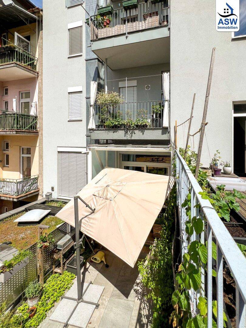 Terrasse mit großem Sonnenschirm und Pflanzen, angrenzend an ein Gebäude mit mehreren Balkonen.