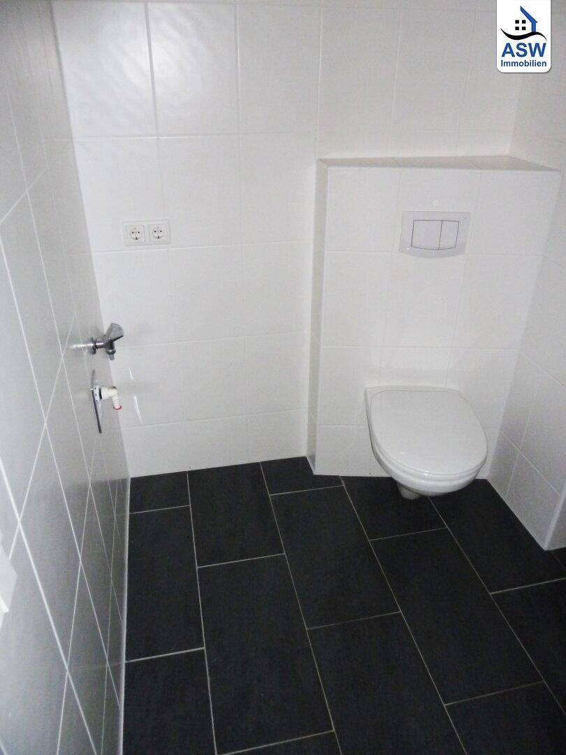 Separates WC mit wandhängender Toilette und dunklen Fliesen, sauber und funktional.