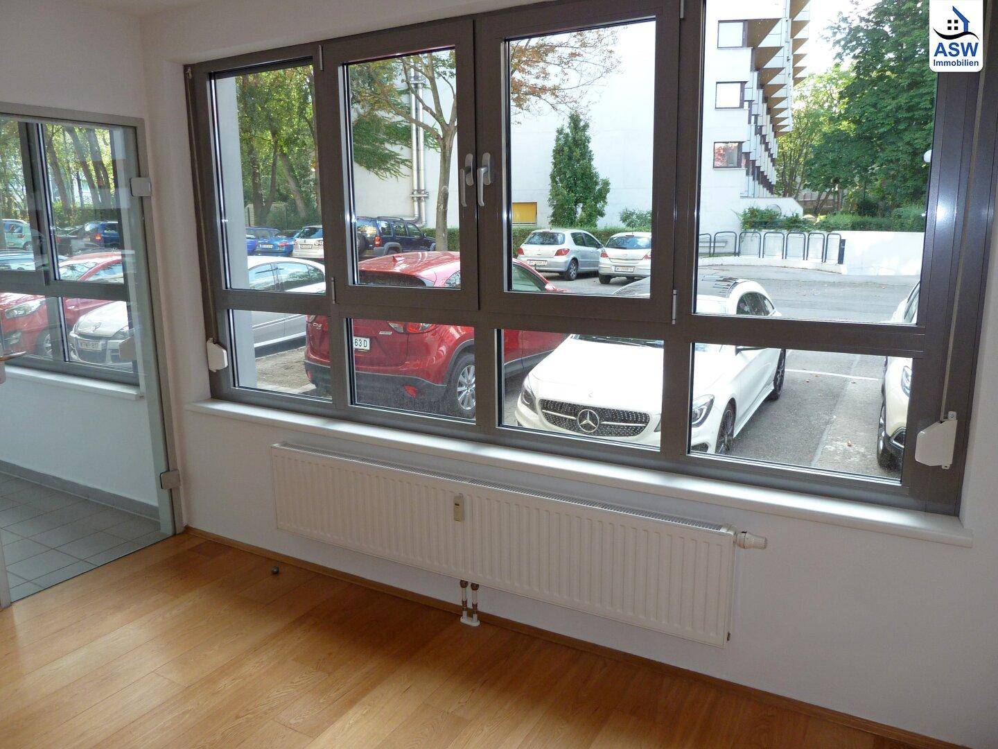 Helles Zimmer mit Holzfußboden und Blick auf den Außenbereich mit parkenden Autos.