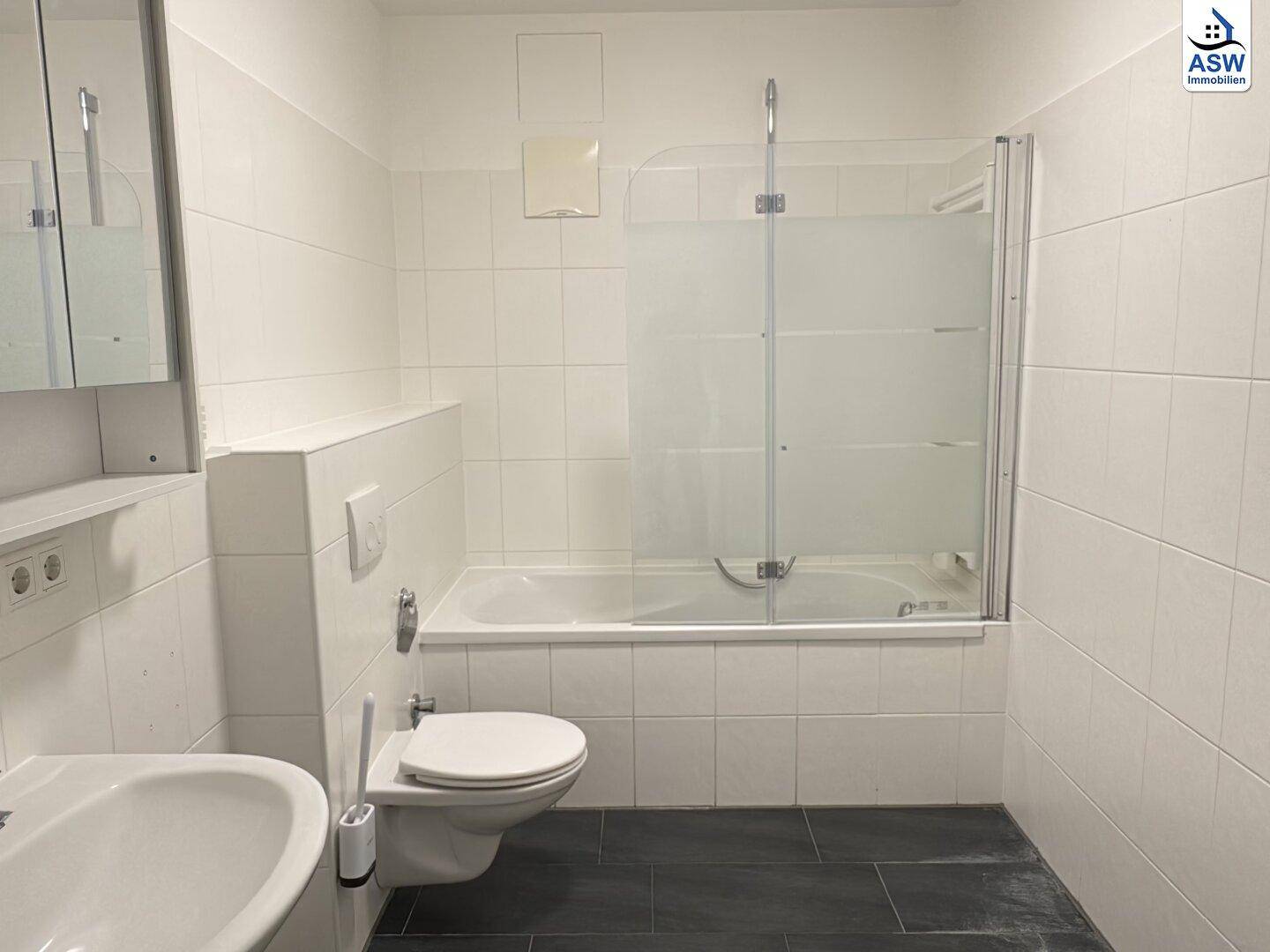 Modernes Badezimmer mit weißen Fliesen, Badewanne mit Duschwand, Toilette und Waschbecken.