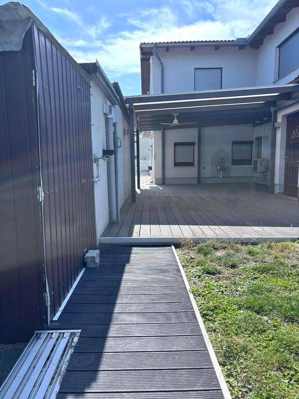 Überdachte Holzterrasse mit angrenzendem Schuppen und Blick auf das Wohnhaus.