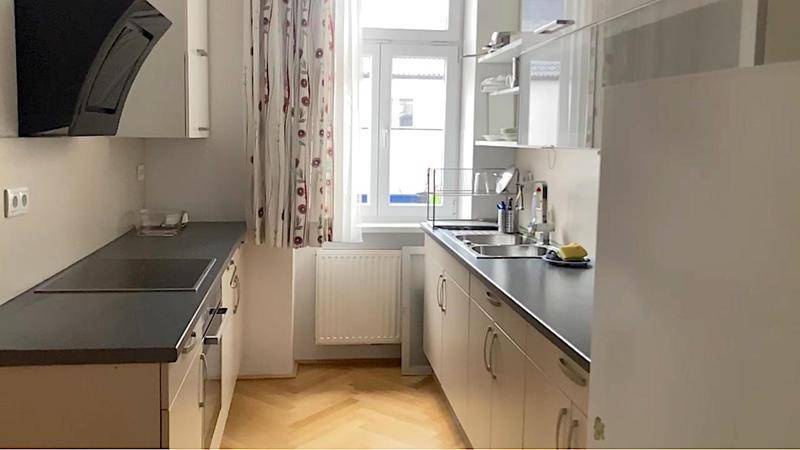 Helle Küche mit beigen Unterschränken, dunkler Arbeitsplatte, Dunstabzugshaube und Fenster.