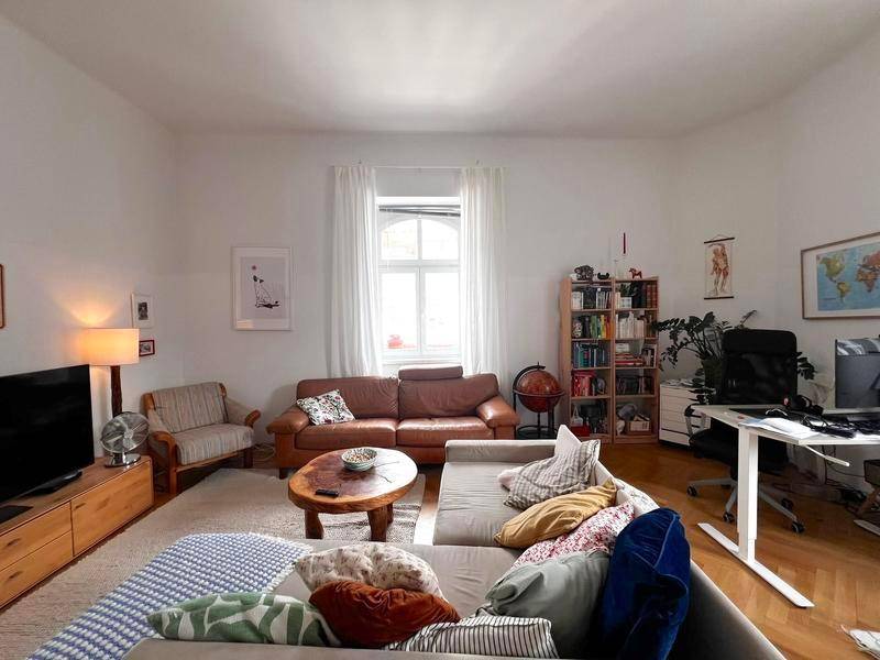 Geräumiges Wohnzimmer mit Sitzbereich, Schreibtisch und Bücherregal, durch große Fenster hell erleuchtet.
