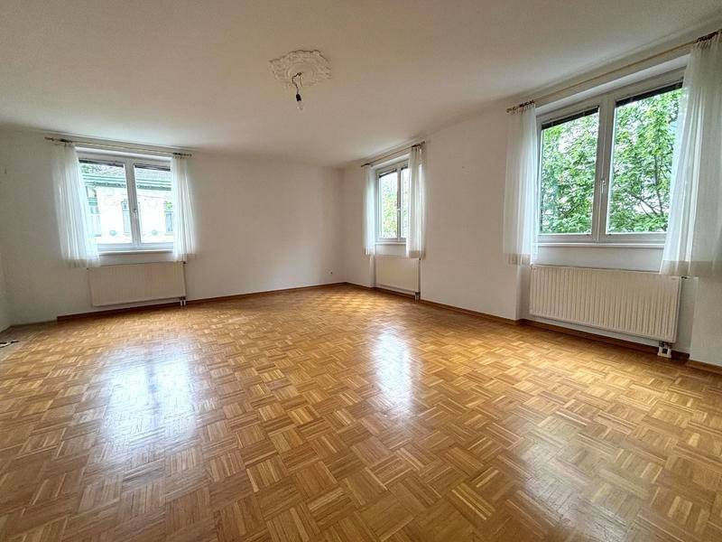 Geräumiger, leerer Raum mit Fischgrätparkett und zwei Fenstern mit Blick ins Grüne.