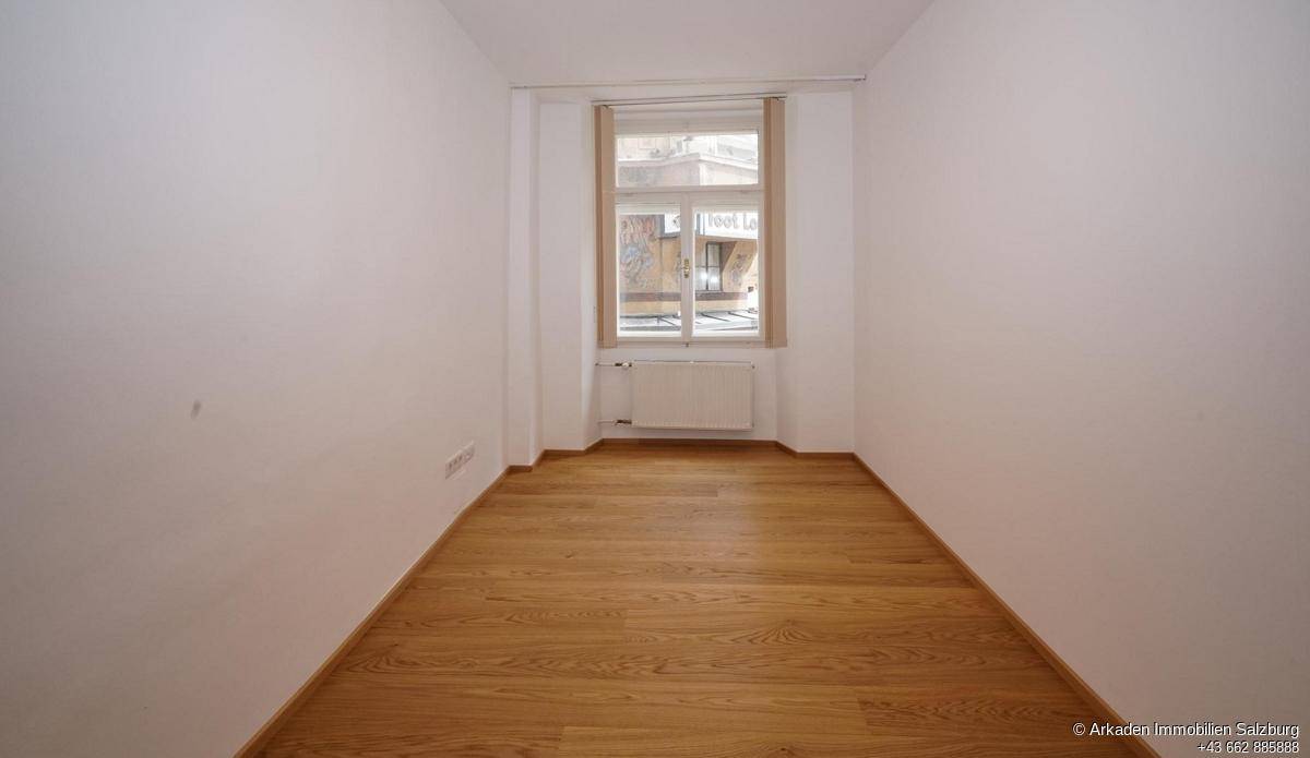 Schmaler, heller Raum mit Holzboden, weißen Wänden und einem Fenster mit Blick auf die Straße.