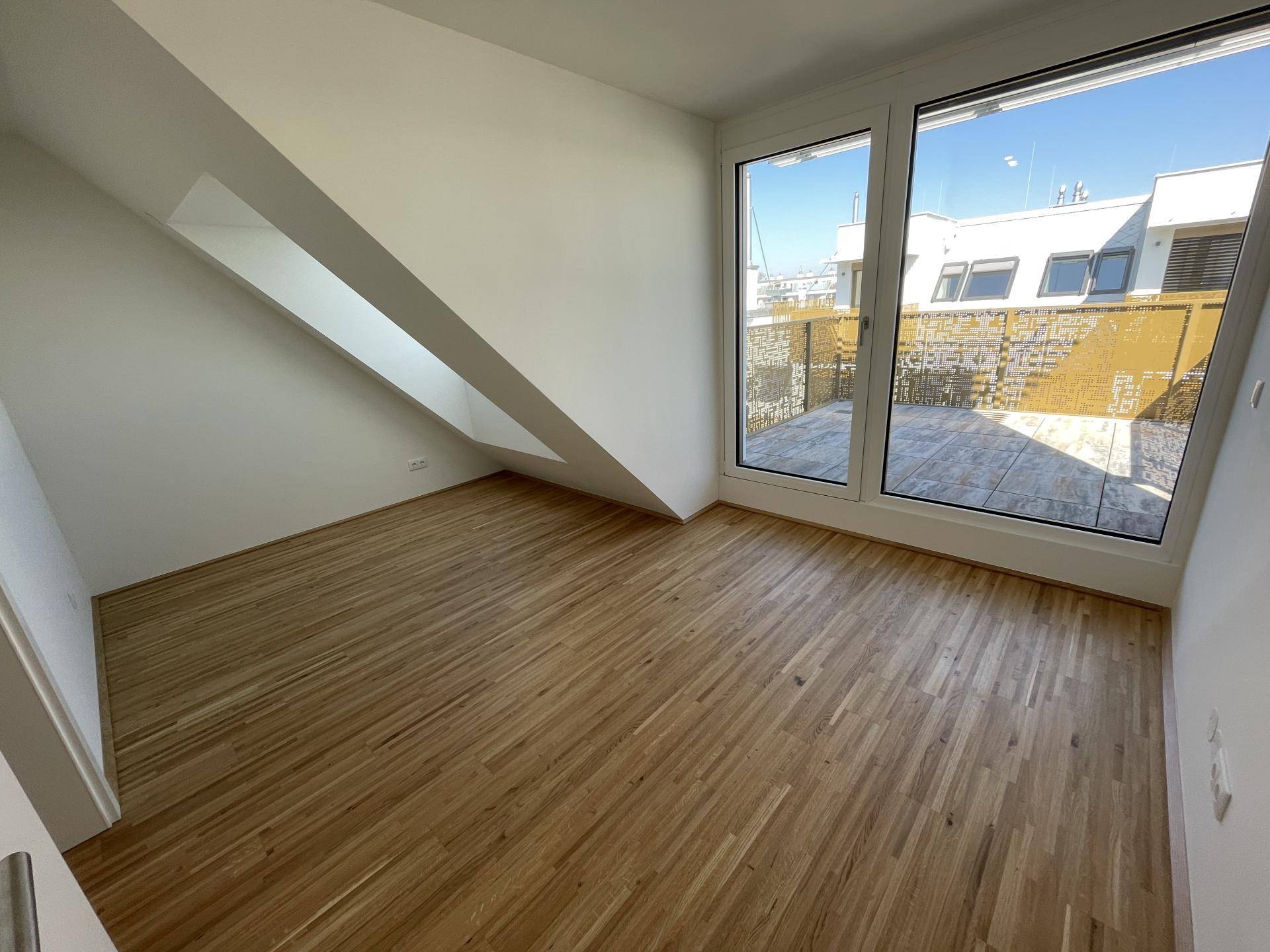 Helles Zimmer mit großen Fenstern, Parkettboden und Zugang zur Außenterrasse.