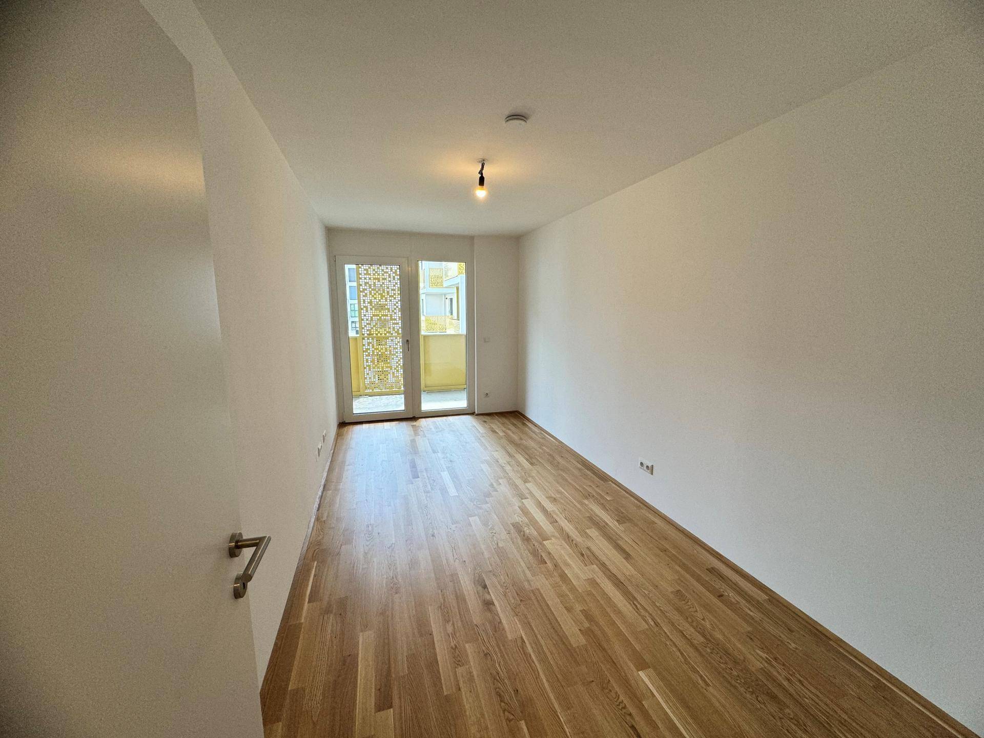 Helles, leeres Zimmer mit Parkettboden und Zugang zu einem Balkon.