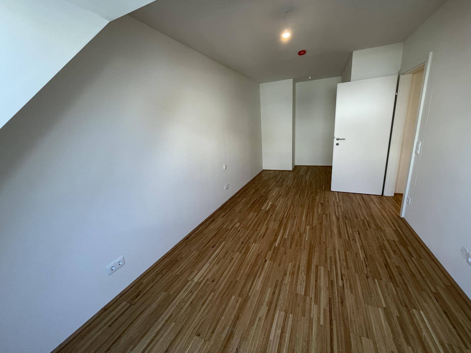 Leeres Zimmer mit Holzboden, weißen Wänden und Dachschrägen, bereit zur Einrichtung.