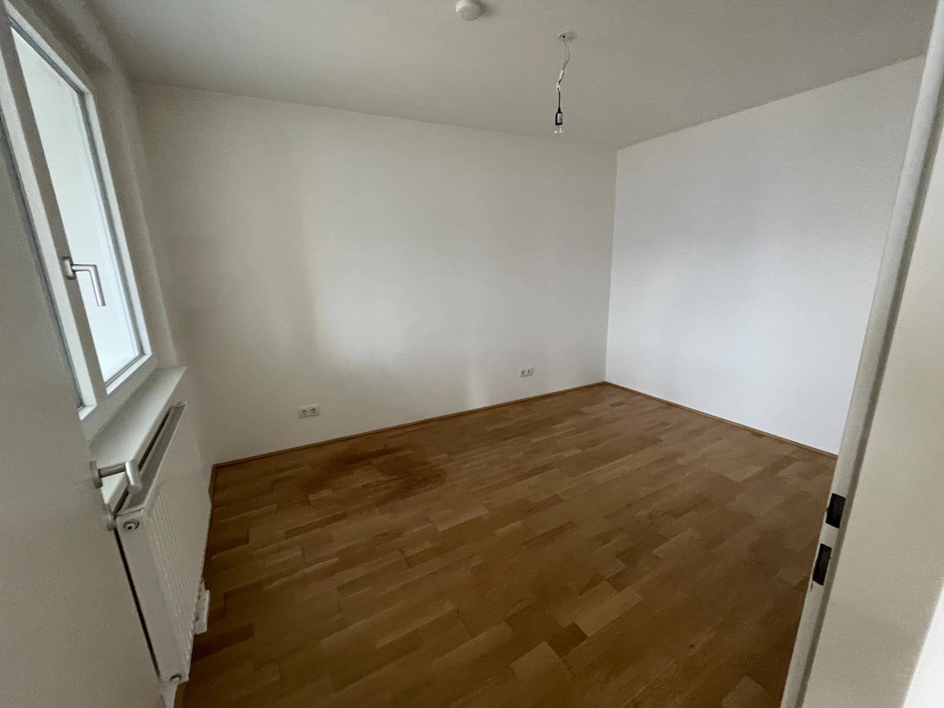 Leeres Schlafzimmer mit Parkettboden, weiß gestrichenen Wänden und einem Fenster.