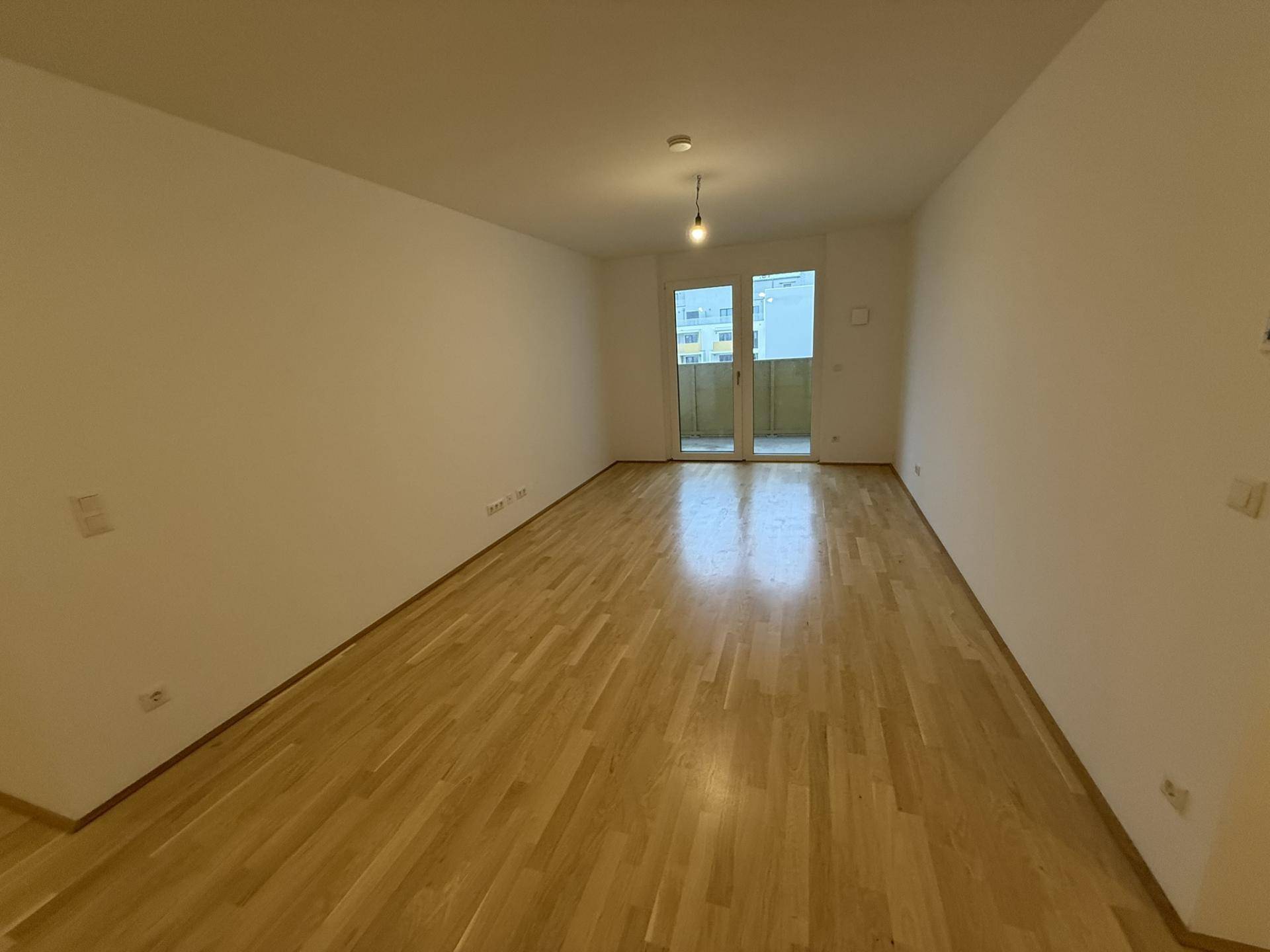 Geräumiges, leeres Zimmer mit Parkettboden und großen Fenstern zum Balkon.