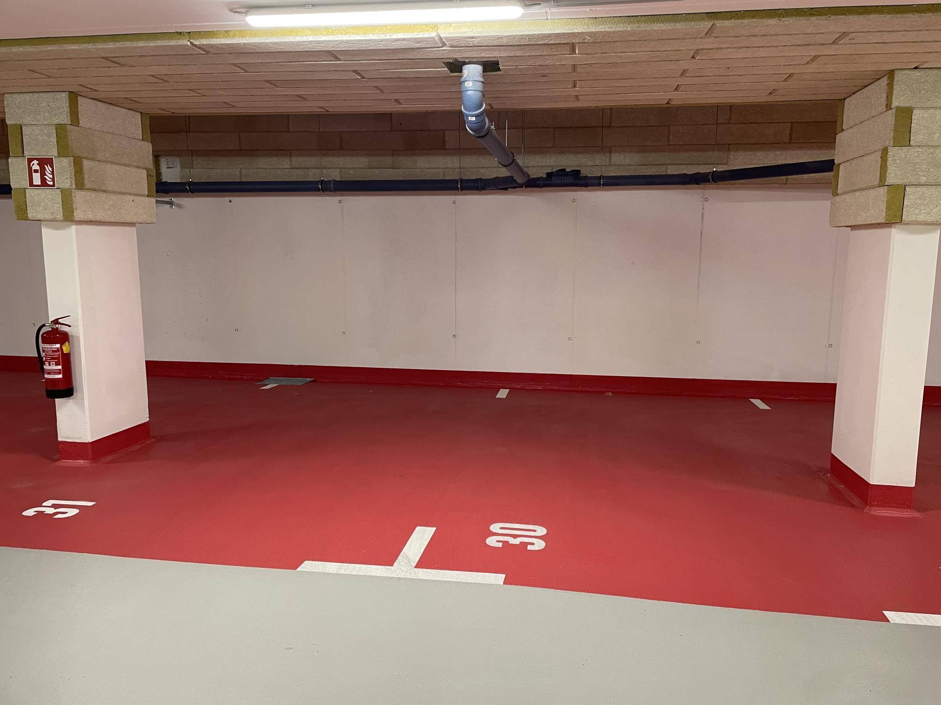 Tiefgaragenbereich mit markierten Stellplätzen, tragenden Säulen und sichtbaren Installationen an der Decke.