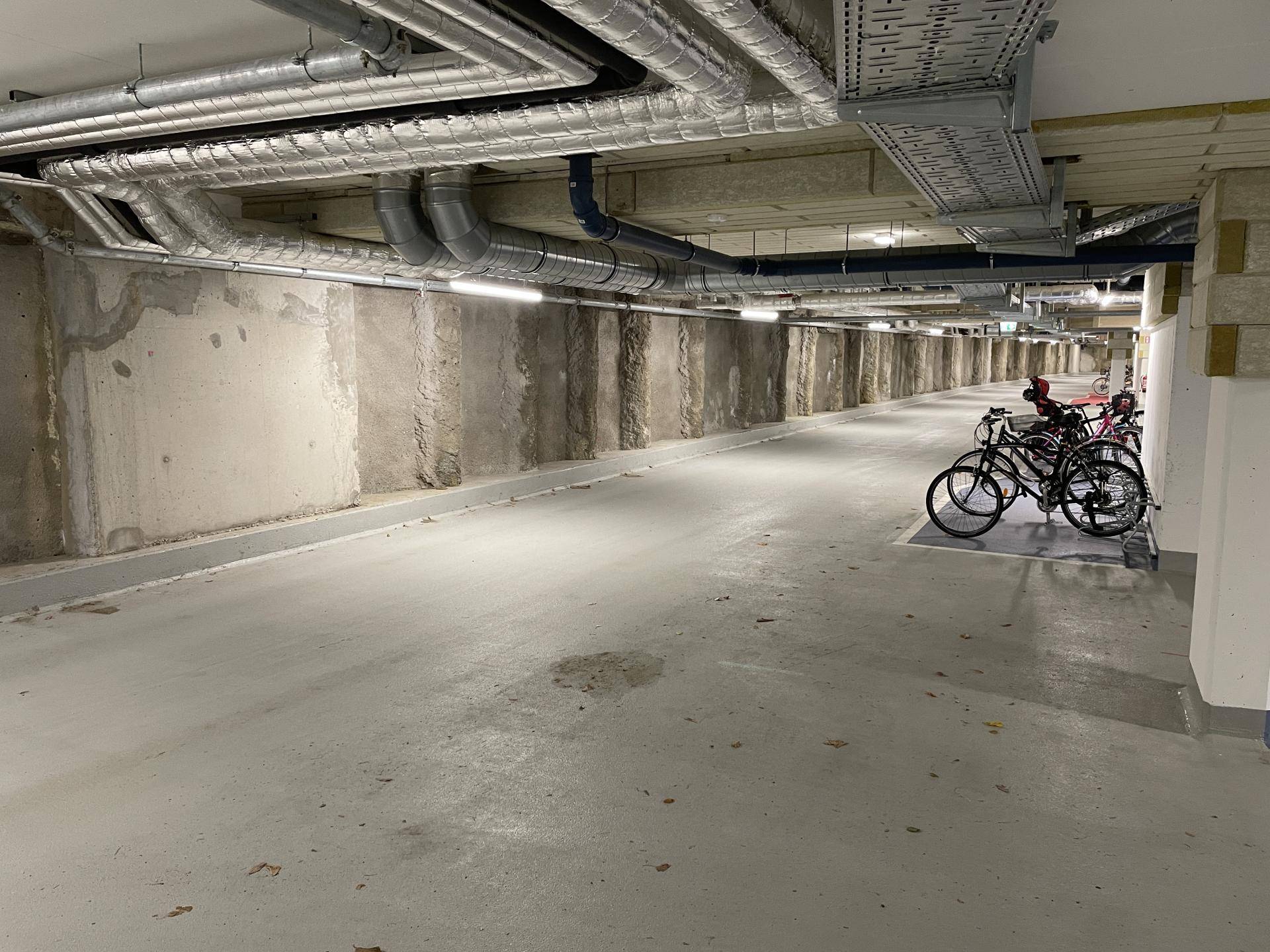 Langer Gang in der Tiefgarage mit sichtbaren Leitungen und abgestellten Fahrrädern.