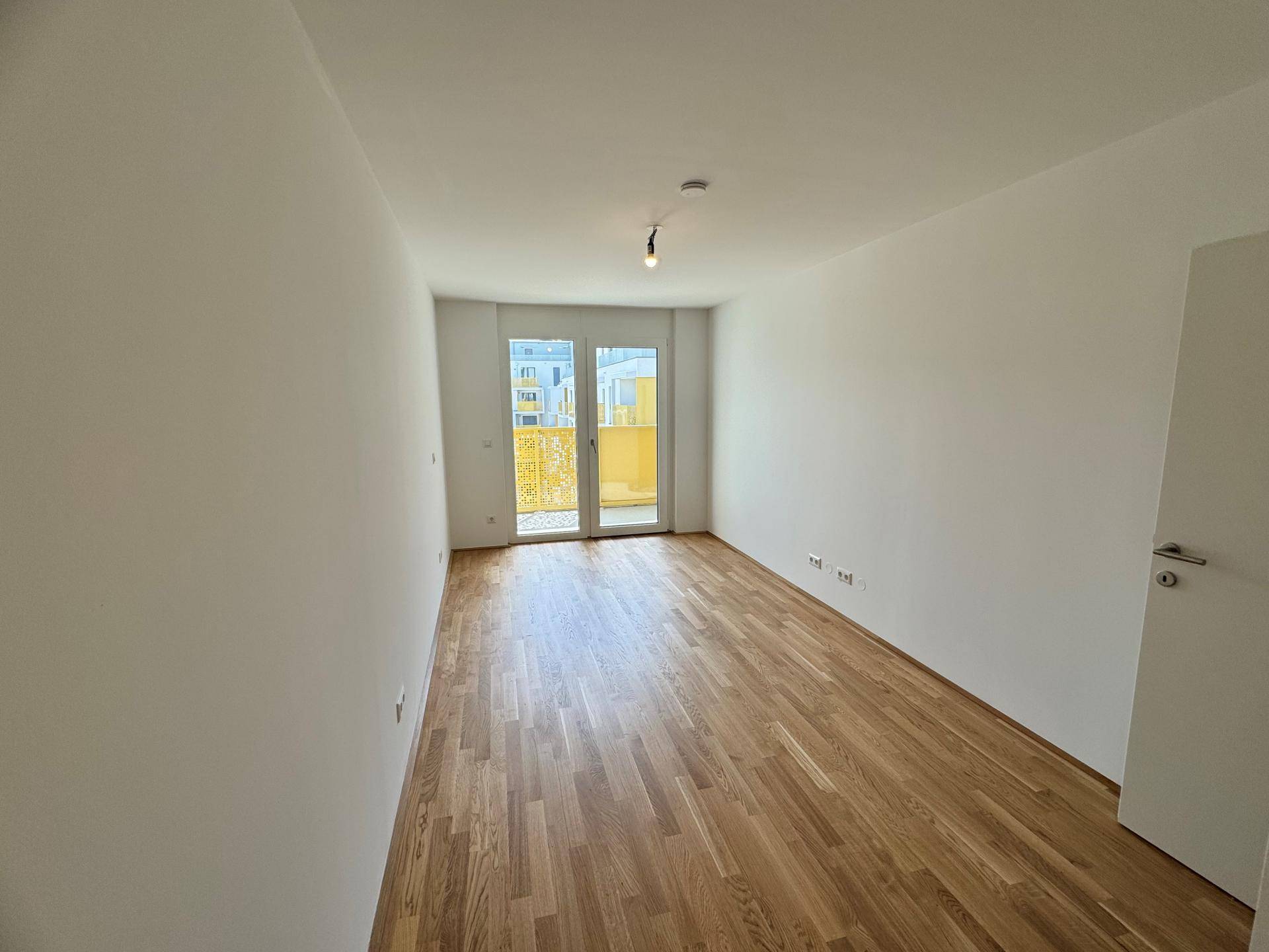 Leeres Schlafzimmer mit Holzboden, weißen Wänden und Zugang zu einem Balkon.