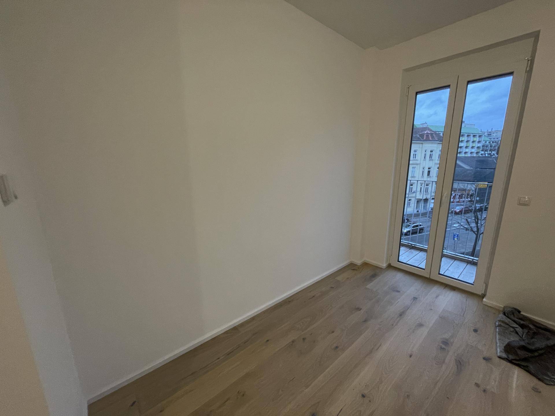 Leeres Zimmer mit Holzboden und Zugang zu einem französischen Balkon, bietet einen Blick auf die Stadt.