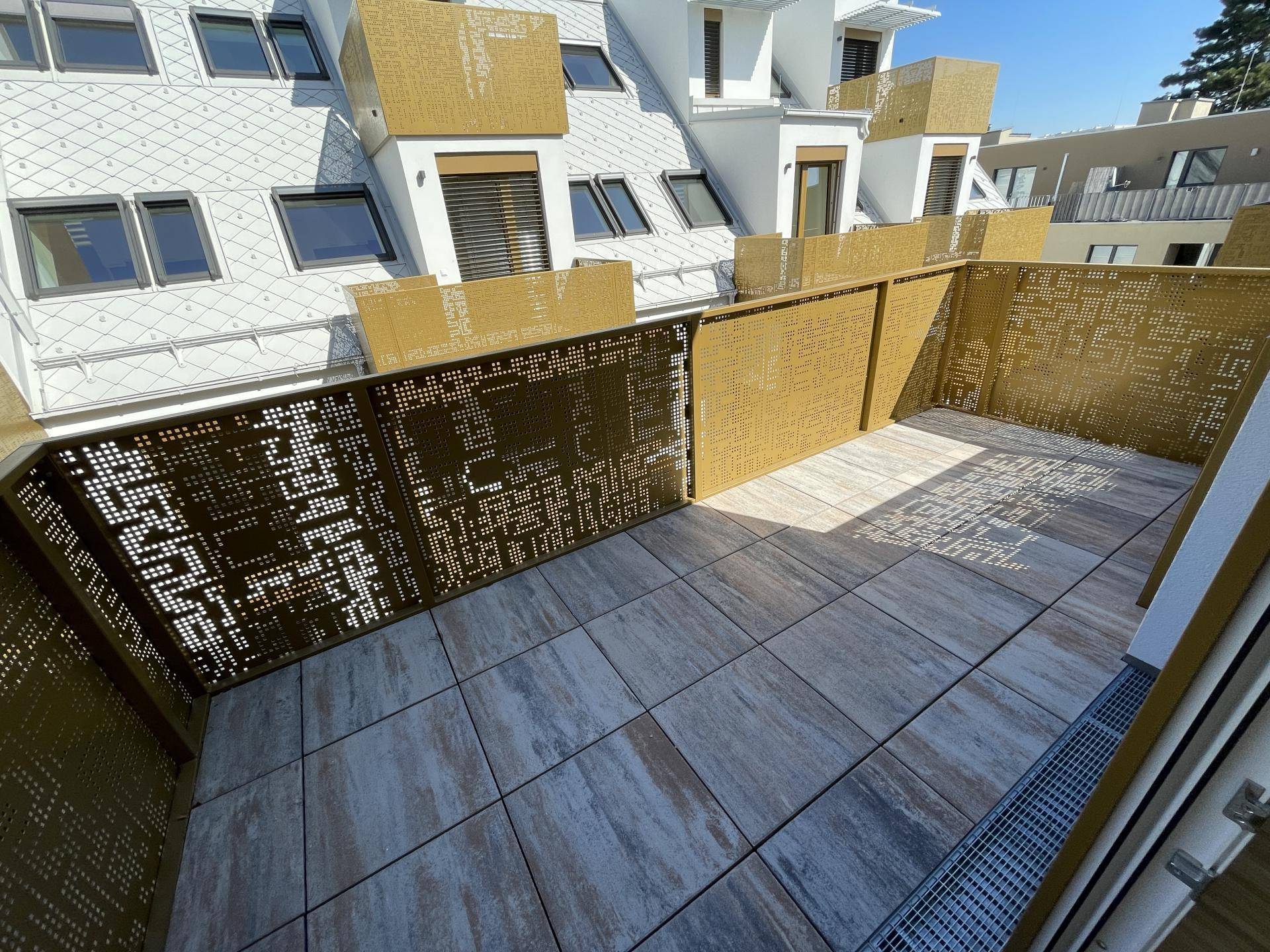 Sonnige Terrasse mit modernen Fliesen und dekorativen Metallgeländern, umgeben von Neubauten.
