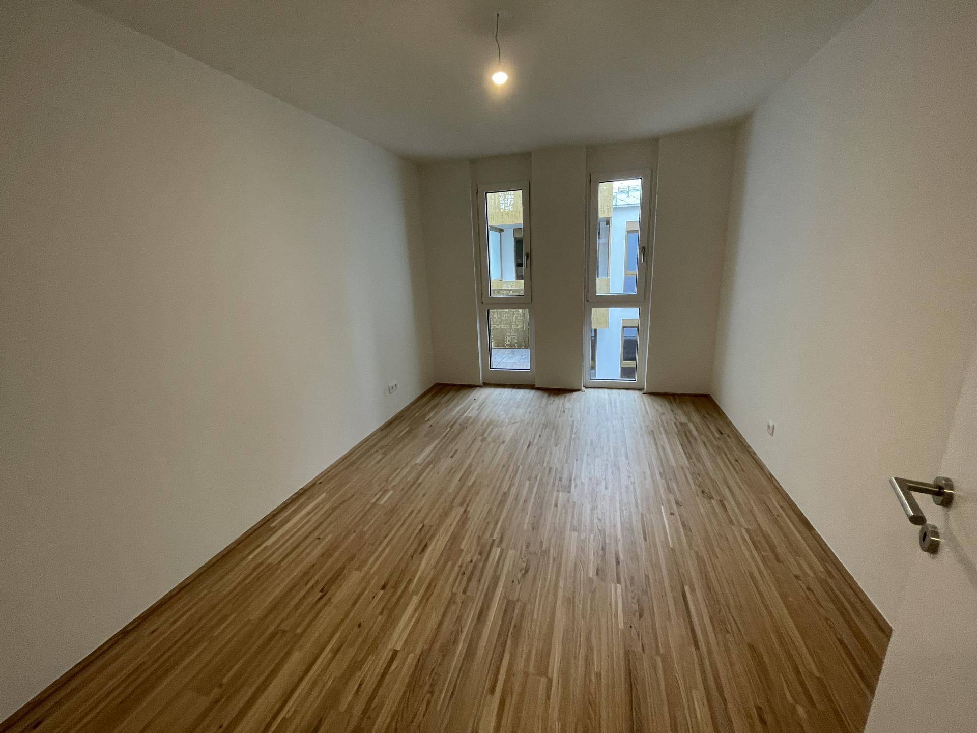 Leeres Schlafzimmer mit hellem Parkettboden und zwei hohen Fenstern.