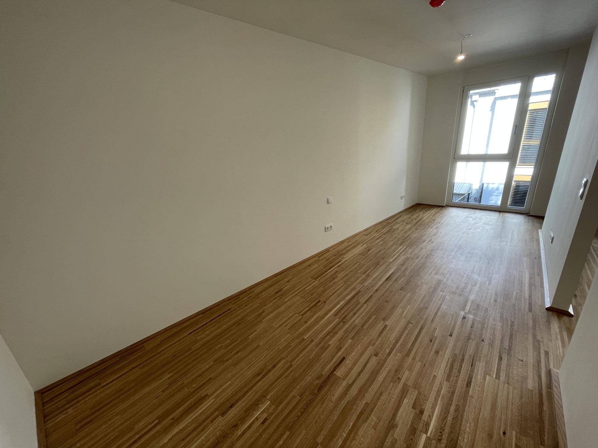 Leerer Raum mit hellem Parkettboden und großem Fenster, das viel Tageslicht hereinlässt.