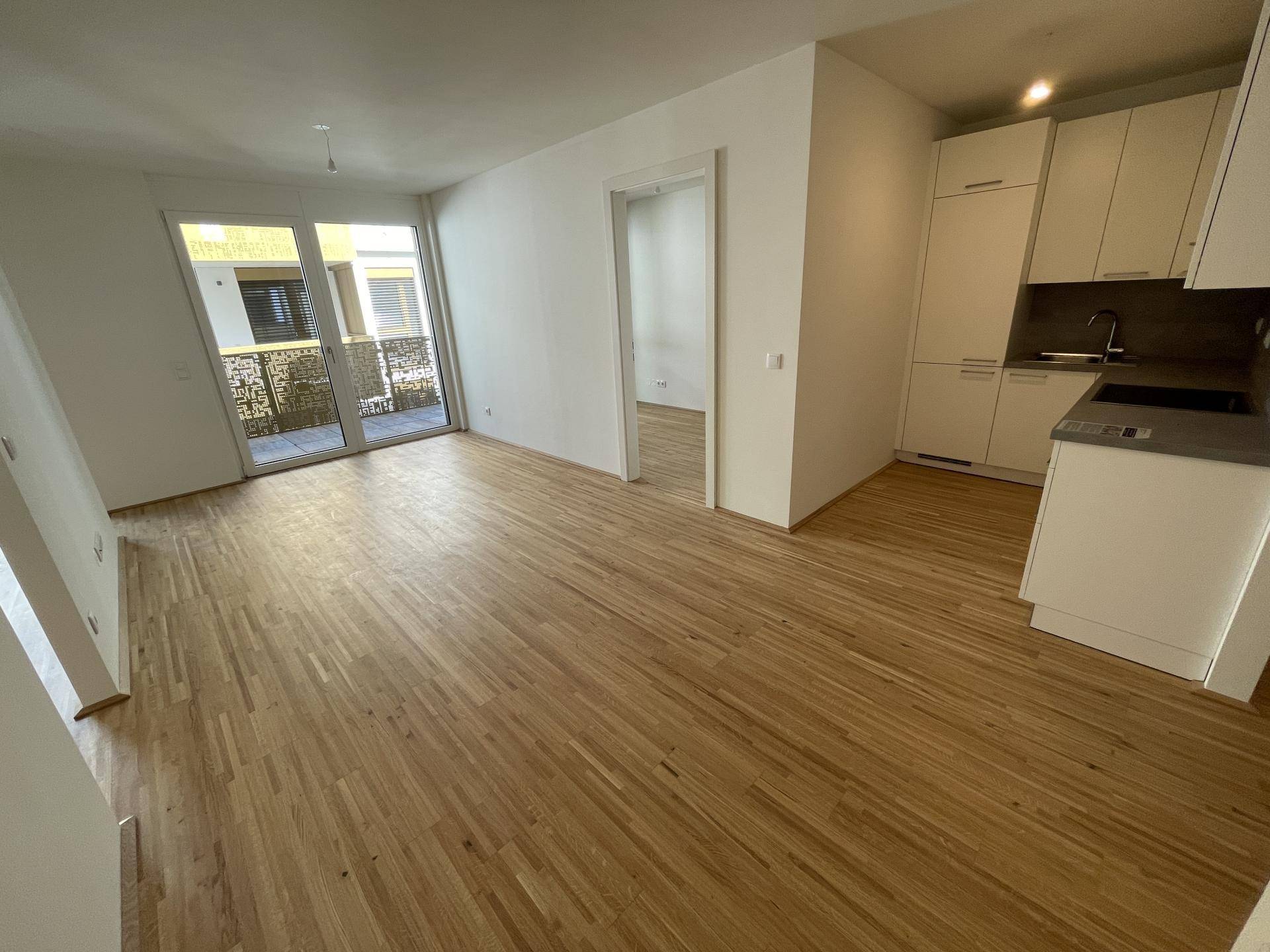 Heller Wohnbereich mit Parkettboden und Zugang zu einem Balkon, angrenzend an eine Küchenzeile.