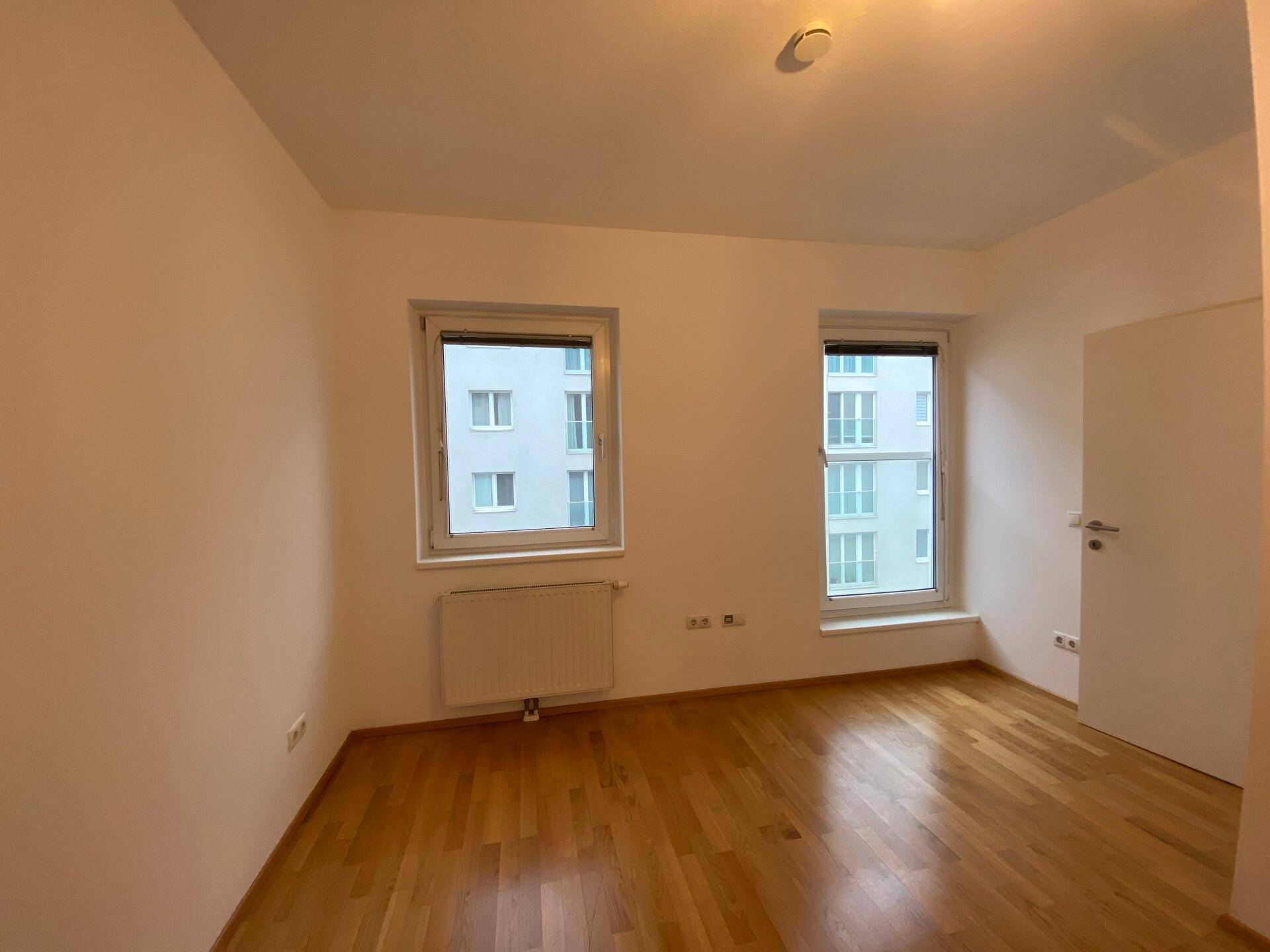 Leeres Schlafzimmer mit Parkettboden und zwei Fenstern, die einen Blick auf die Umgebung bieten.