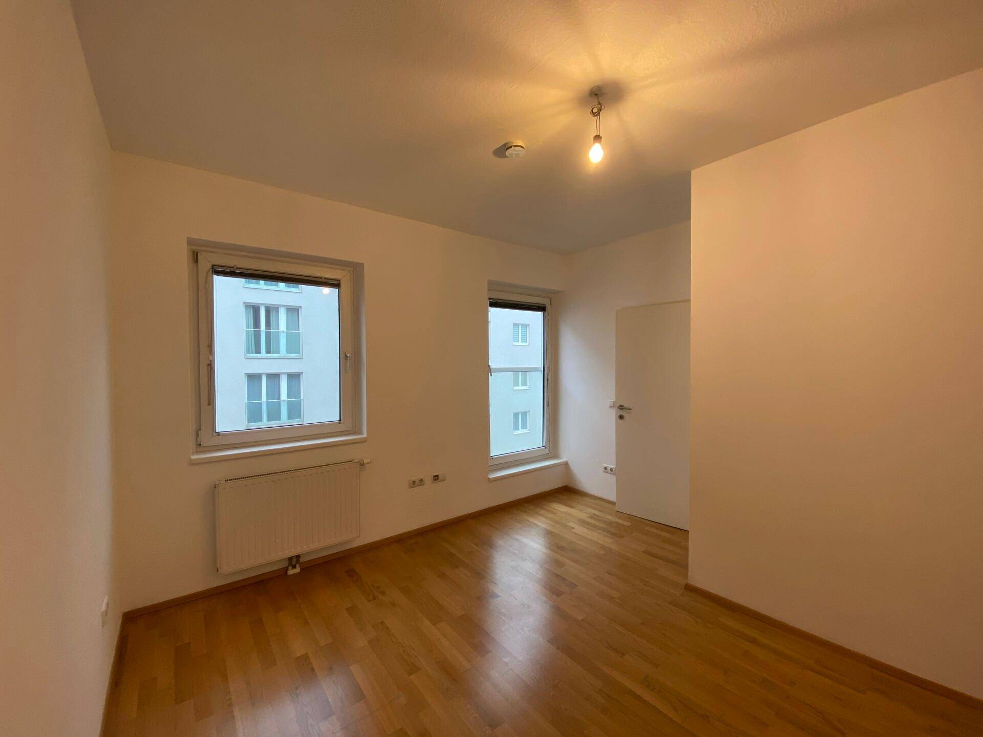 Helles Schlafzimmer mit Parkettboden und zwei Fenstern, die viel Tageslicht hereinlassen.