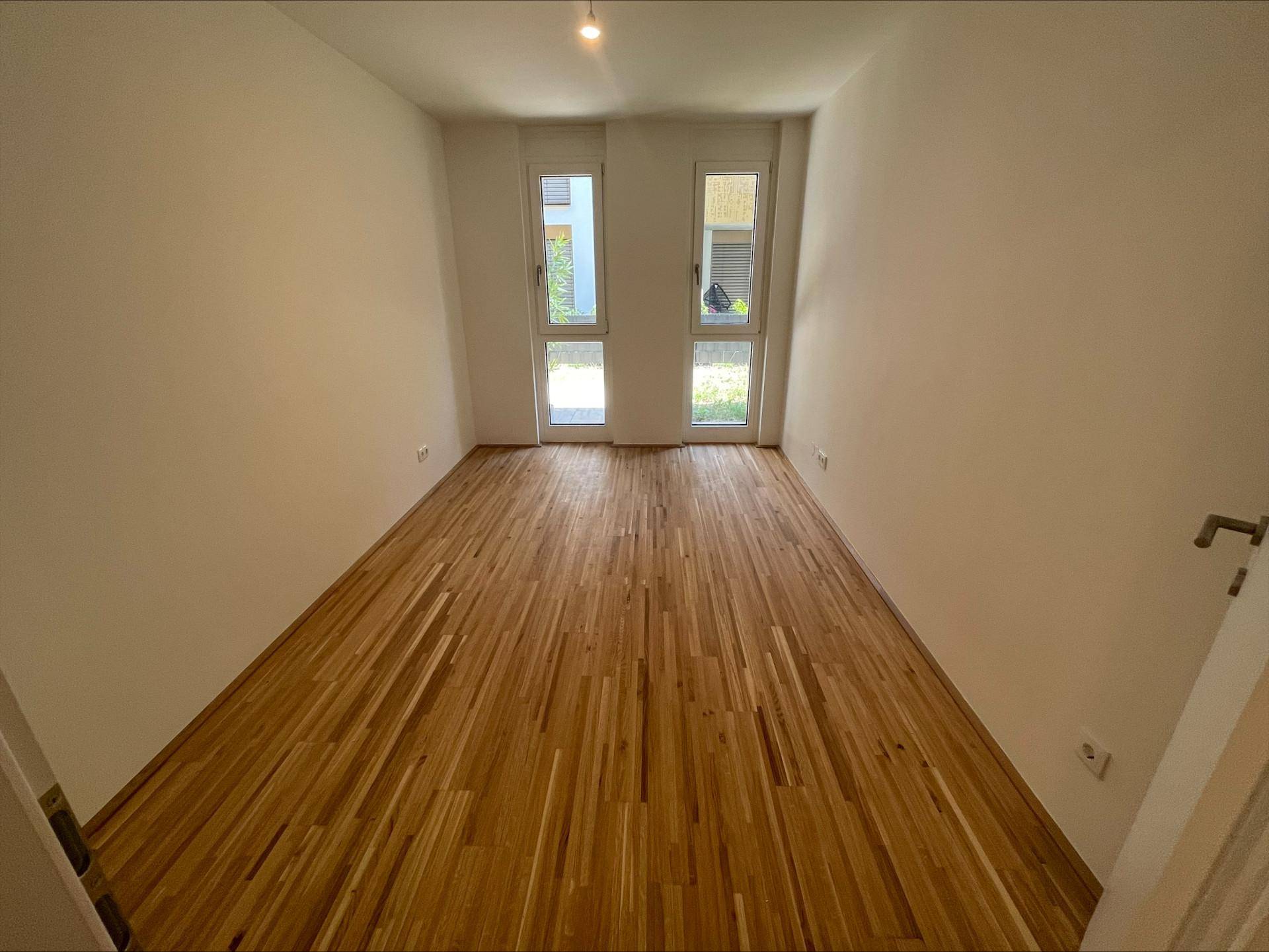 Leeres Zimmer mit Parkettboden und zwei Fenstern, ideal als Schlaf- oder Arbeitszimmer.