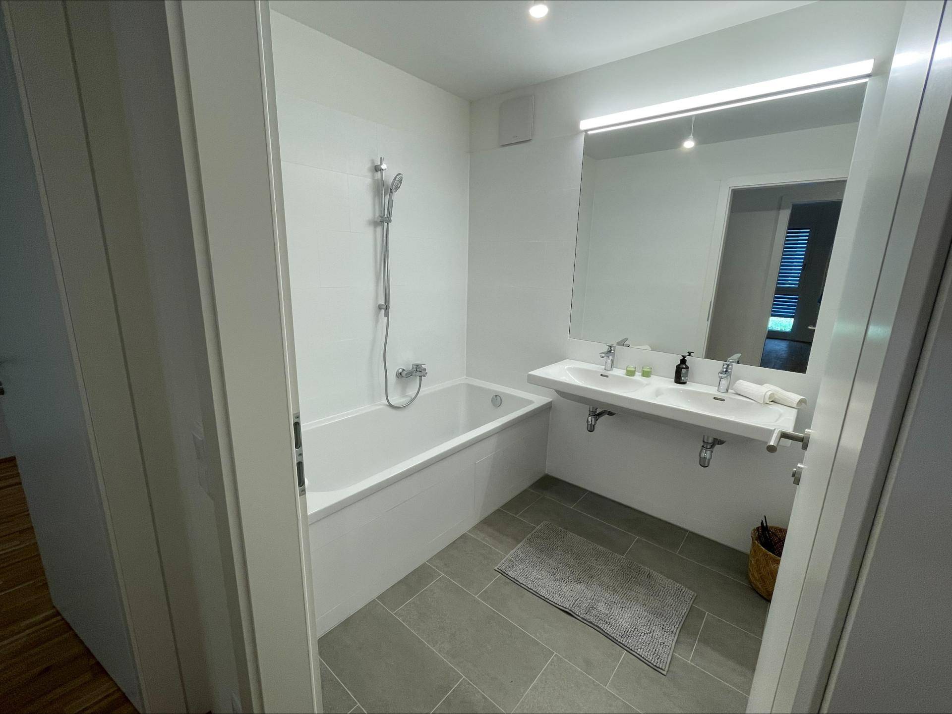 Modernes Badezimmer mit Badewanne, Duschfunktion und Doppelwaschbecken auf Fliesenboden.