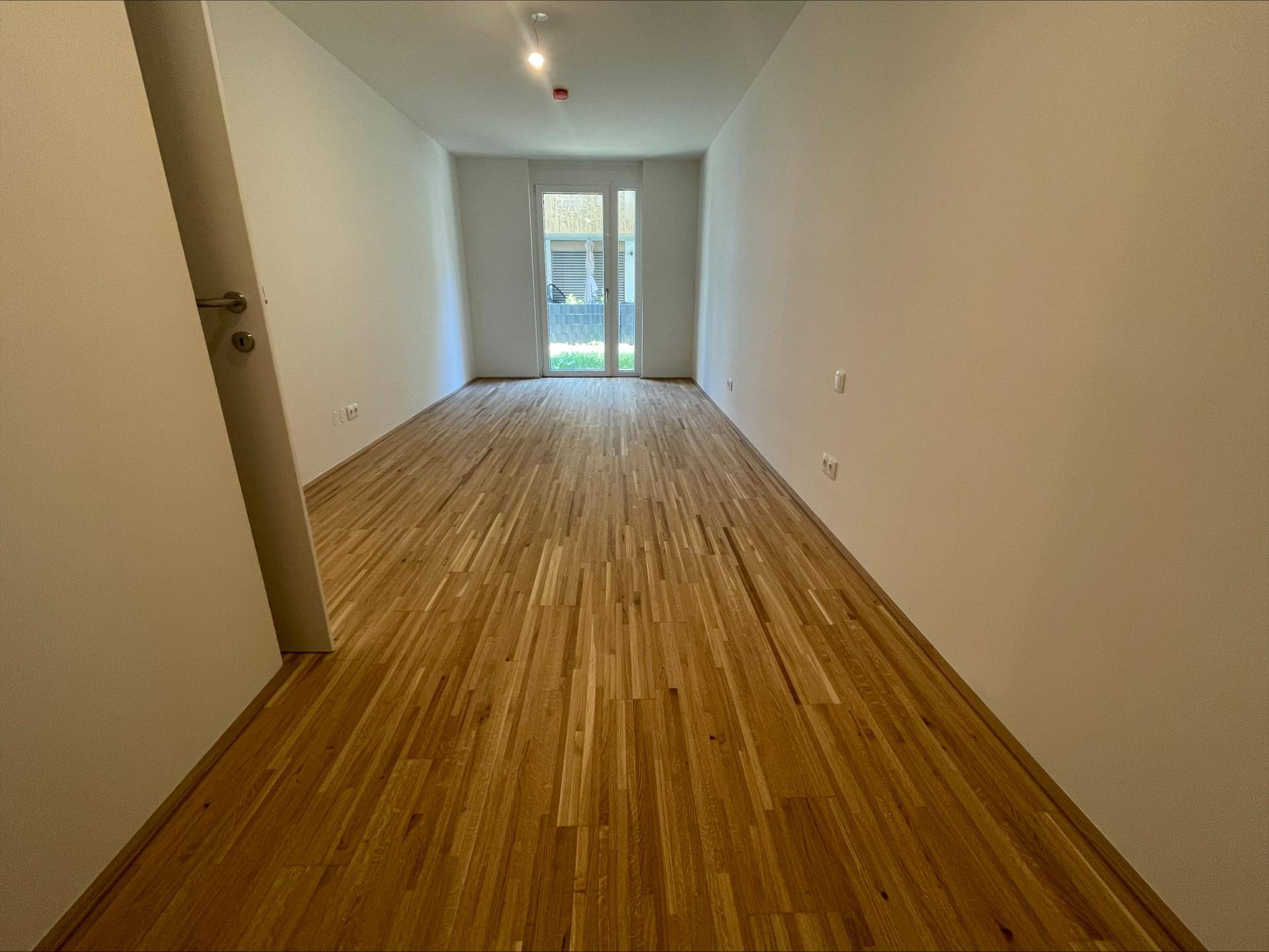 Helles Zimmer mit Holzboden und Zugang zu einem Außenbereich durch eine Glastür.