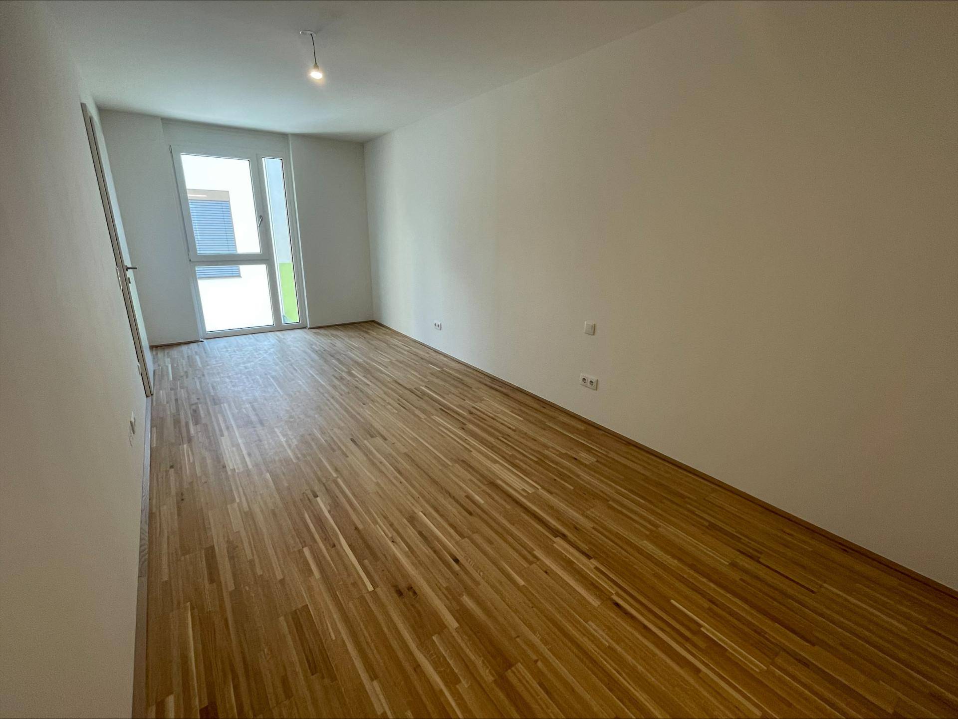 Helles, unmöbliertes Zimmer mit Holzboden und großem Fenster mit Zugang nach draußen.
